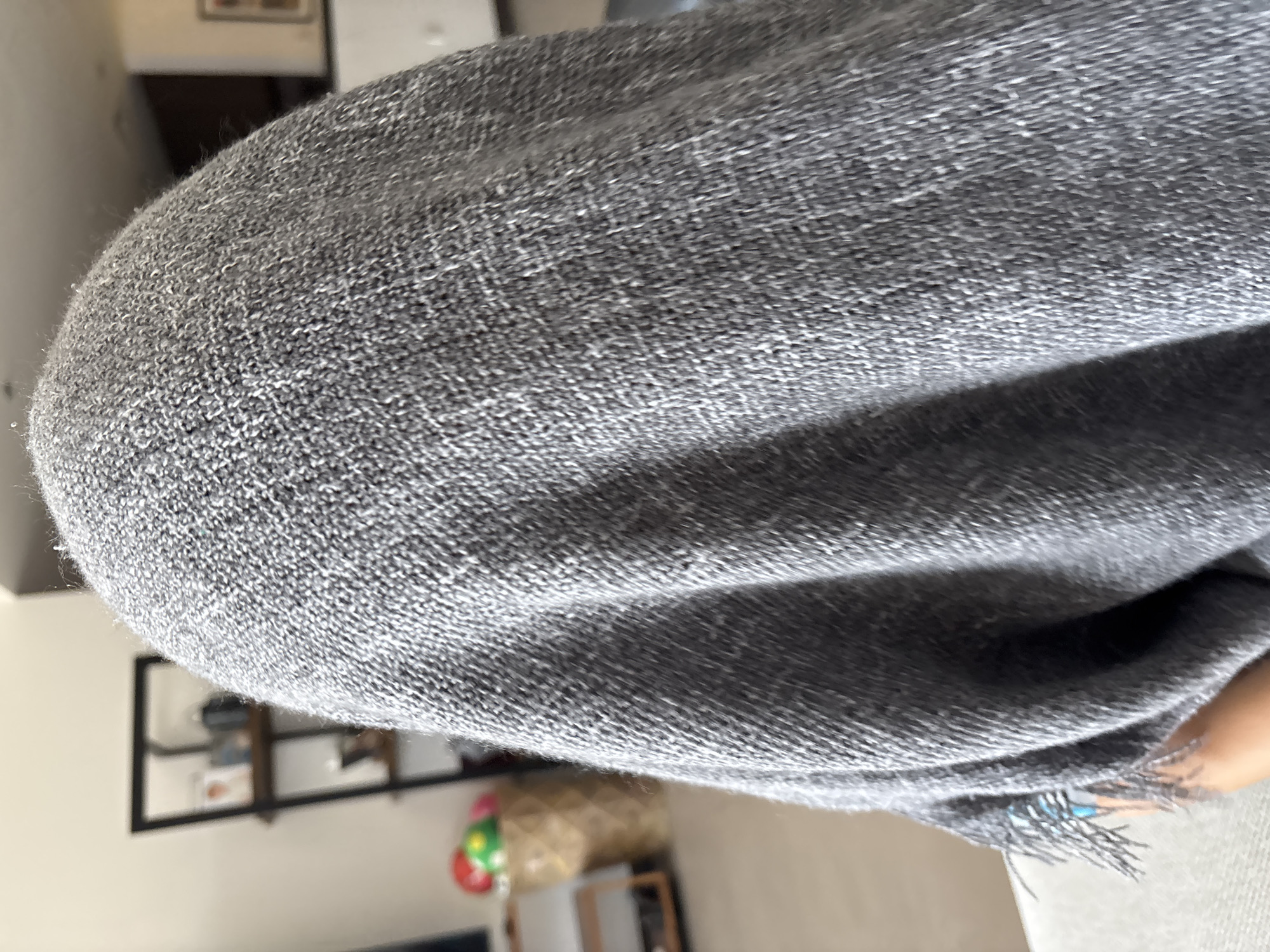 A close-up, over-the-shoulder shot shows the back of a person's head covered by a gray knit blanket or shawl. The person is sitting on a light-colored couch. In the background, out of focus, is a living room setting with a shelf unit, a basket filled with colorful balls, and a framed picture on the wall. The image is taken indoors, with natural light suggesting it is daytime. There is no indication of the specific location within the city of Dubai, United Arab Emirates.