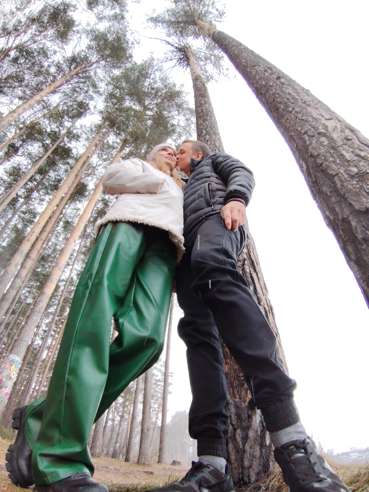 In a low-angle shot, a couple stands in a forest in Chaikovsky, Russia. The woman, on the left, wears a white jacket with a furry trim and glossy green pants. The man, on the right, wears a black puffer jacket and black pants. He leans against a tree, kissing the woman on the cheek. Tall pine trees surround them, stretching upwards toward the cloudy sky.