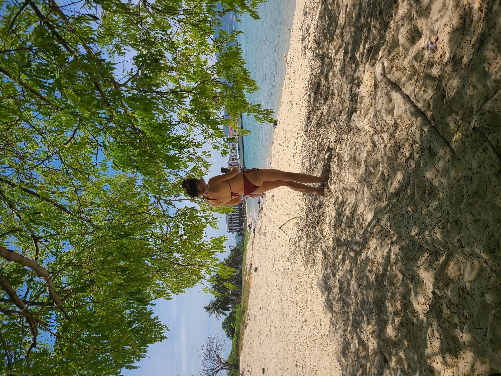 A woman in a maroon bikini stands on a sandy beach, facing away from the camera and looking at her phone. She is positioned under the shade of a tree with green foliage, its branches extending into the top of the frame. The beach is light-colored sand, with scattered leaves and small debris. In the background, the turquoise ocean meets a hazy blue sky. Further in the distance, along the coastline, are palm trees, a few buildings, and what appears to be a small pier or jetty. The scene is consistent with a beach in Nha Trang, Vietnam, a popular coastal city known for its sandy beaches and tropical scenery.