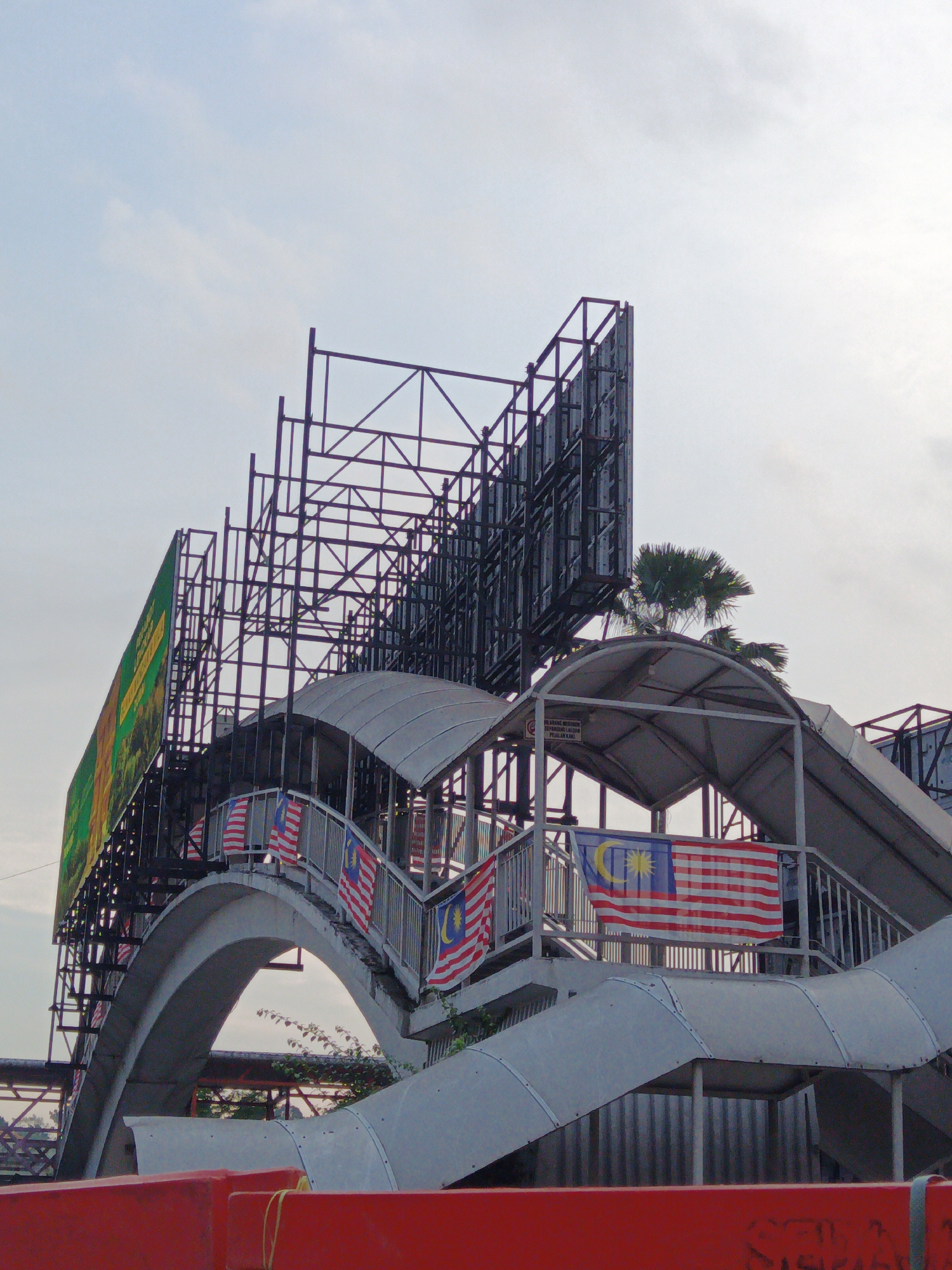 This is a photograph of a pedestrian bridge and billboard structure under a cloudy sky in Kuala Lumpur, Malaysia. The bridge, made of concrete and metal, has an arched ramp and stairs. Several Malaysian flags are displayed on the railings of the bridge. Behind the bridge, a large metal framework supports a billboard that is partially covered by a green banner. Palm trees are visible in the background, suggesting a tropical urban environment. In the foreground, red traffic barriers are visible. The image appears to be taken during daylight.