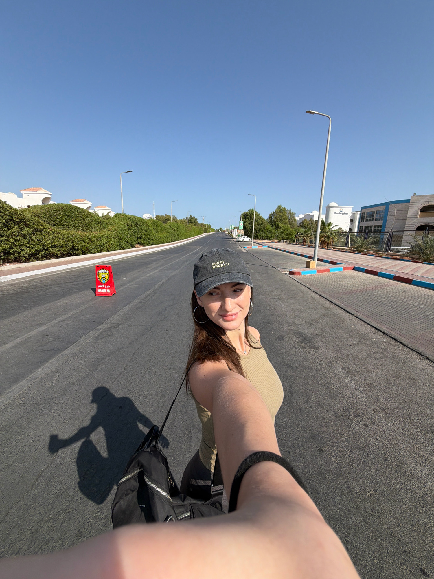 A woman is standing on an empty asphalt road in Sharm El Sheikh, Egypt, holding a camera to take a selfie. She is wearing a grey baseball cap that says "SUPER happy :)" and a sleeveless olive-green top. A black backpack is slung over her shoulder. Her shadow, cast by the bright sun, is visible on the road. The road is lined with manicured green hedges and white buildings with orange tiled roofs on the left side, and a sidewalk with decorative colored borders on the right. In the distance, there are more buildings, including one with a blue and white facade. A red "TRAFFIC" sign with the words "NO PARKING" is visible on the left side of the road. The sky is clear and blue.