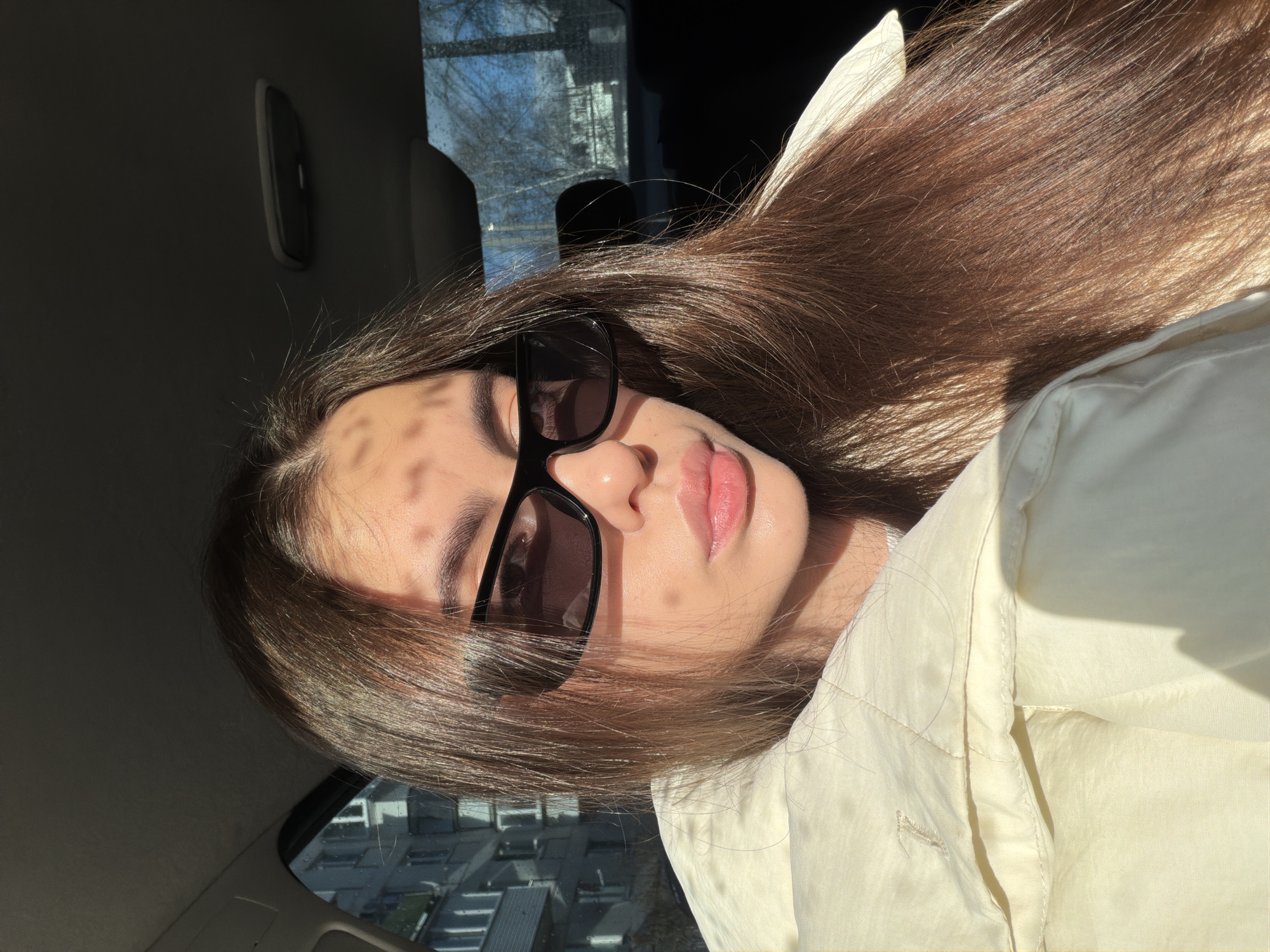 A young woman with long, dark hair is seen in the backseat of a car. She is wearing sunglasses and a light-colored jacket. The sun is shining through what appears to be a building. The exact location in Chaikovsky, Russia, cannot be determined.