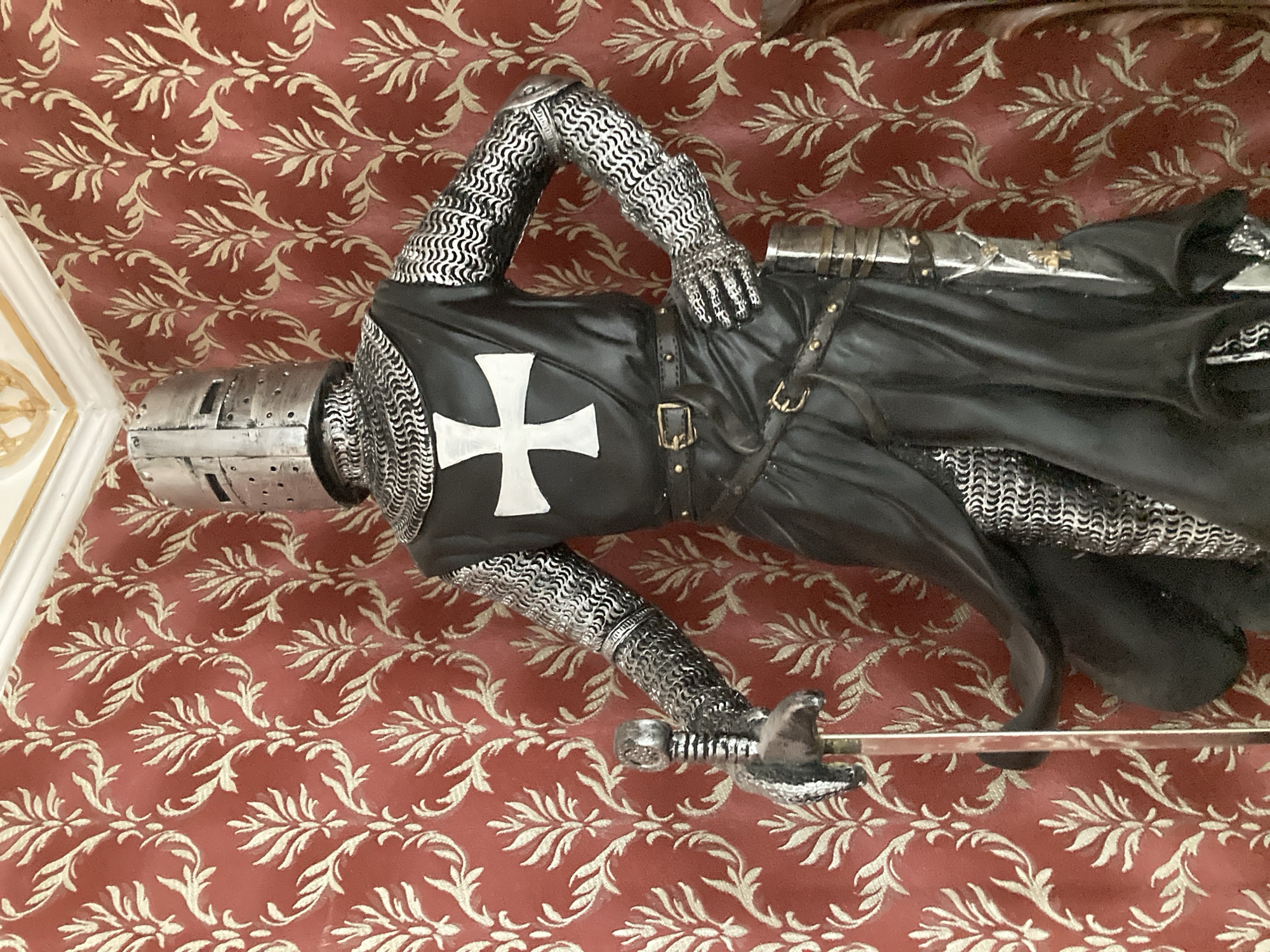 A statue of a knight in full armor stands against a red damask wallpaper. The knight wears a silver helmet with a visor, a black tunic with a white cross on the chest, and chainmail on the arms and legs. A sword is held in the knight's right hand, and a scabbard is visible on the left side of the waist. The background features a repeating pattern of stylized leaves in a lighter color against the red. The scene is lit from above, creating shadows and highlighting the metallic textures of the armor.