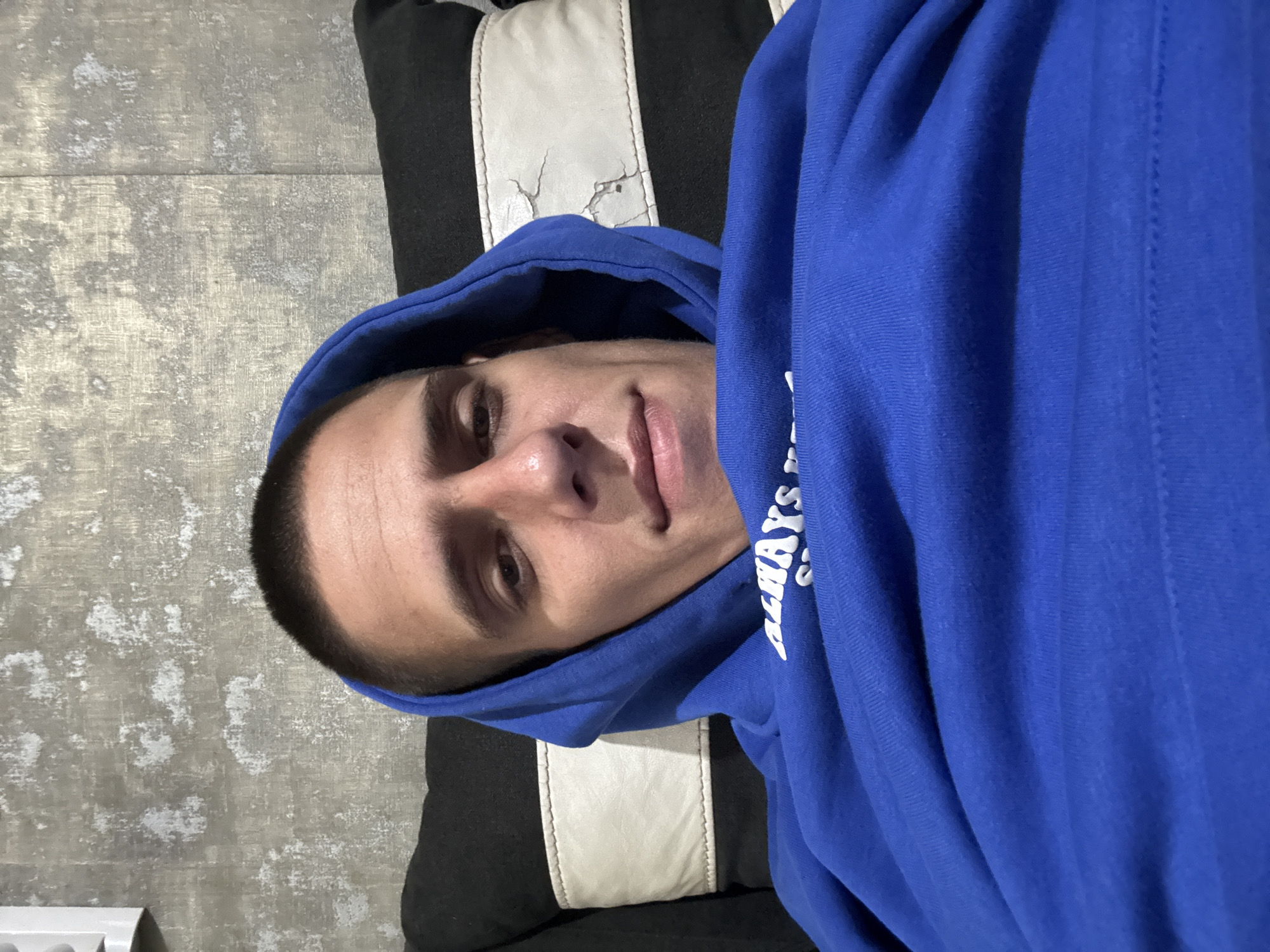 A close-up, self-taken photograph shows a young adult male with short dark hair and a close-shaved haircut. He is wearing a bright blue hoodie with the hood pulled up around his face. The background consists of a textured gray wall and a pillow with black and white striped sections, one of which shows signs of wear and tear. The lighting appears to be artificial, casting soft shadows on the man's face and the surrounding objects. The man's expression is neutral, and he is looking directly at the camera. There are no discernible landmarks or contextual clues to determine the location within the city of Sofia, Bulgaria, or any specific urban environment. The image is solely focused on the individual and his immediate surroundings.