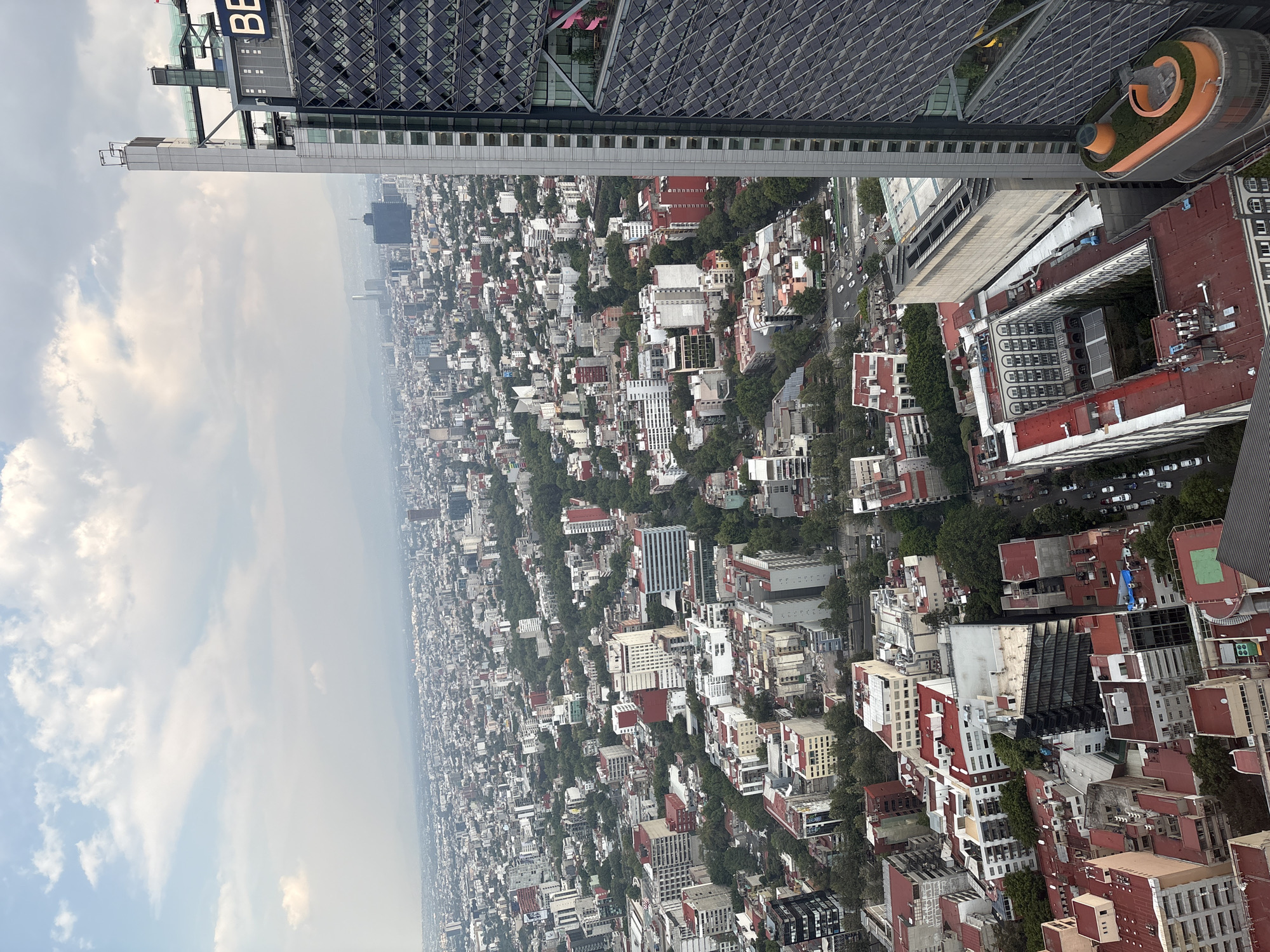 The image provides a high-angle, panoramic view of a vast urban landscape, characteristic of a major metropolis like Mexico City, under a partly cloudy daytime sky. The perspective is from a significant height, looking out over an extensive sprawl of buildings that extends to a hazy horizon.
In the foreground and midground, a dense array of architectural styles is visible. A prominent, tall grey skyscraper on the left side clearly displays the text "BBVA" near its top. Adjacent to it, another modern building features a distinctive dark facade with an intricate, geometric pattern. A pedestrian skybridge connects two sections of buildings. Below these high-rises, the city unfolds with a mix of residential and commercial structures, many with red-tiled roofs, densely packed and interspersed with numerous green trees and parks lining streets and filling courtyards. One building features a unique rooftop garden with an orange, curved structural element. Roads with visible vehicular traffic weave through the urban fabric.
The sky above is blue with scattered white clouds, transitioning to a lighter, somewhat hazy tone towards the distant city skyline, which fades into the atmospheric haze. No specific human or animal activities are discernible due to the elevated vantage point. The scene captures the immense scale and density of a bustling city during daylight hours, with a mix of modern and traditional architecture amidst significant urban greenery.