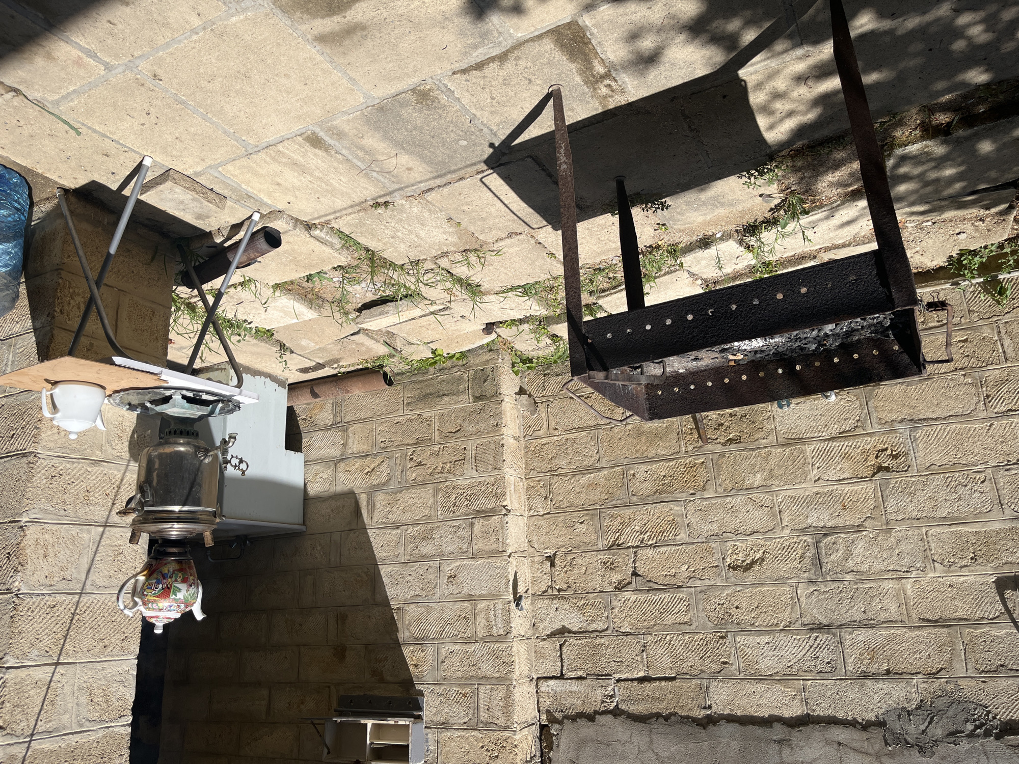 This is an outdoor scene in Sumqayit, Azerbaijan, featuring a brick-walled courtyard. A black metal barbecue grill, containing ashes, is positioned on the left side of the frame. To the right, a silver samovar with a decorative teapot on top is present, next to a small white teapot on a portable folding table. The ground is paved with large square tiles and scattered with some vegetation. Sunlight casts shadows across the scene, indicating a sunny day. The background consists of a stone wall and a white structure.