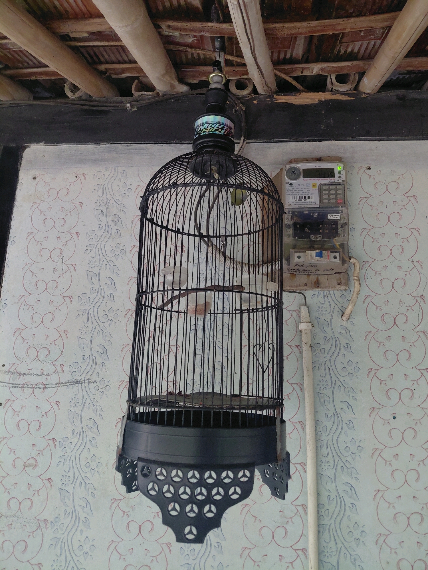 The image shows a black birdcage hanging from a wooden beam, suspended in front of a patterned wall. An electric meter is mounted on the wall to the right of the birdcage. The birdcage is ornate, with decorative metalwork at the bottom and a smaller cage or perch inside. A small bird, possibly yellow or light green, is visible within the cage. The wall has a repeating floral pattern in shades of pink and blue. The ceiling structure, visible at the top of the image, is made of wooden beams and appears to be part of a traditional or rustic construction. The overall scene suggests an outdoor or semi-outdoor setting, possibly a porch or veranda. The presence of the electric meter indicates a residential or commercial building. The image is taken from a slightly upward angle, looking towards the ceiling.