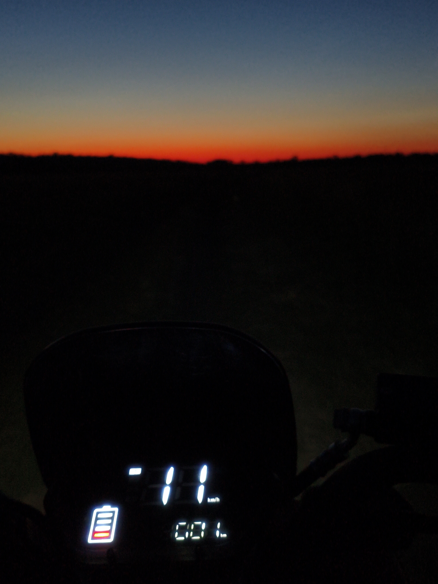 This is a close-up, low-angle, outdoor photograph of a motorcycle's illuminated dashboard at dusk. The sky displays a gradient of colors, transitioning from dark blue at the top to a vibrant orange-red band near the horizon. Below the horizon, the landscape is rendered in deep shadow, with only the silhouettes of what appear to be fields or low vegetation visible. The motorcycle's dashboard, prominently featured in the foreground, is black and glossy, reflecting the dark environment. It displays several illuminated digital readouts. On the left is a battery indicator showing approximately one-quarter charge, represented by a red bar within a white battery outline. To the right of the battery indicator, the numbers "-11" are displayed in bright white, followed by "km/h" on the right. Below these numbers, "00 L" is displayed, also in bright white. The overall impression is one of a journey beginning or ending in the twilight.