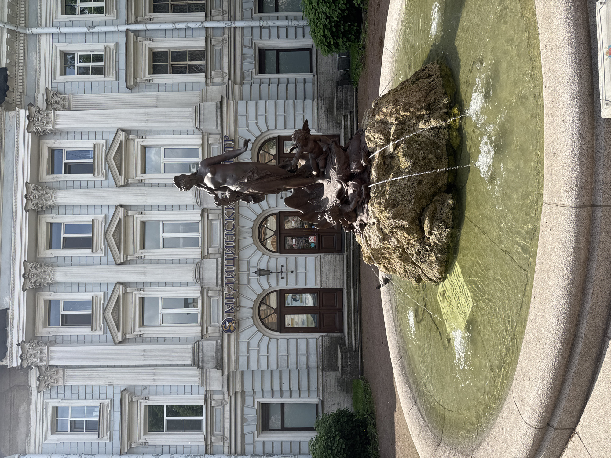 In Saint Petersburg, Russia, a bronze fountain sculpture stands before a light blue classical building adorned with white columns and arched doorways. The sculpture depicts a female figure standing on a shell-like base, with cherubic figures around her. Water sprays from the rock formation supporting the figures and falls into the circular basin of the fountain, which has a rough-textured stone rim. The building features numerous windows with white frames and intricate architectural detailing. In the background, green bushes and trees are visible. The sky is clear and bright, indicating a sunny day.