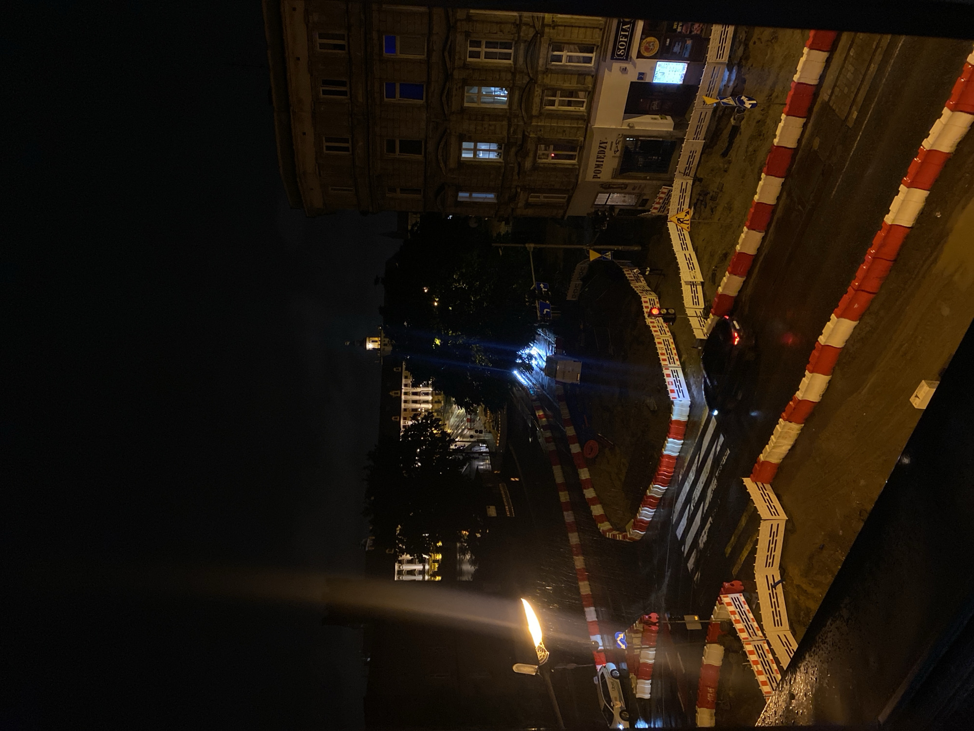 This is a nighttime aerial view of a street scene in Wrocław, Poland, likely during ongoing road construction. The street is wet, suggesting recent rain. Red and white barriers delineate various sections of the road, indicating closed areas and detours. A street lamp illuminates part of the road, casting a bright glow. Several vehicles are visible, including a white car with "Uber" branding, a dark sedan, and a white utility vehicle. Buildings line the street; one on the right is a multi-story stone structure with illuminated windows, displaying signs for "POMIĘDZY" and "SOFIA." In the background, more buildings and trees are silhouetted against the dark sky, with some lights visible. The overall impression is one of a quiet, somewhat damp urban environment at night, disrupted by construction activities.