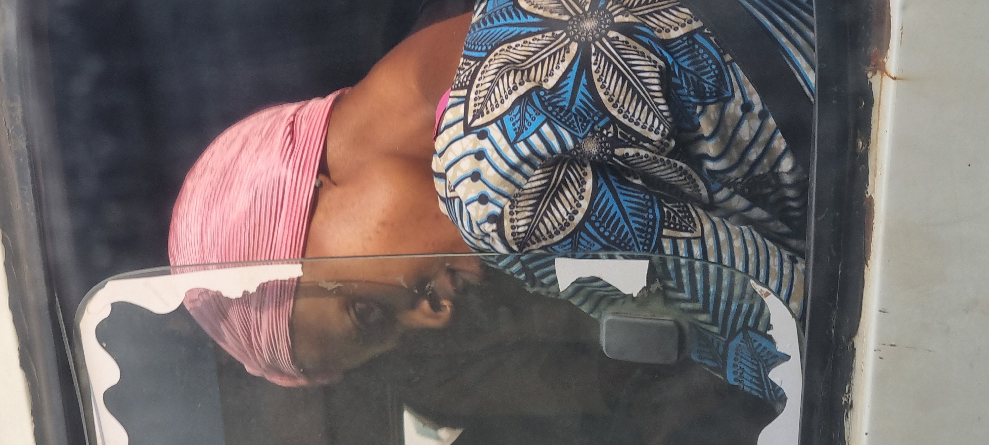A woman wearing a pink headscarf and a blue and white patterned garment is seen through a cracked window of a vehicle. Her reflection is visible in the glass, showing her face turned to the left. The scene appears to be in Otta, Nigeria. The vehicle's interior is dimly lit, contrasting with the brighter outdoor light. The window is broken with some white tape or paper adhered to it. The woman's body is angled away from the camera, with her back and shoulder visible. The pattern on her garment is intricate with floral motifs and geometric shapes. The exterior of the vehicle shows signs of wear, with rust spots visible on the white frame.