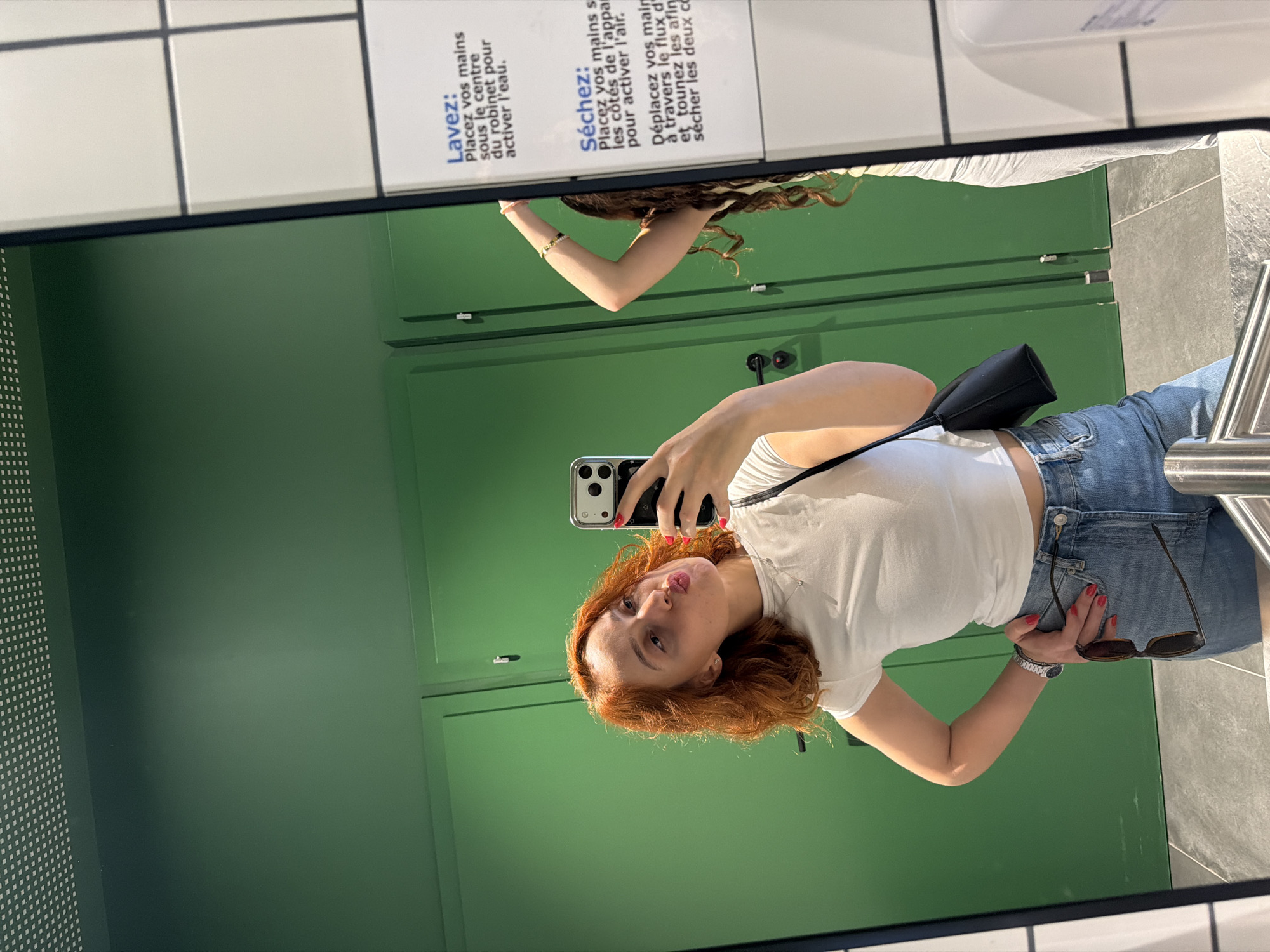 A woman with auburn hair is taking a selfie in a restroom with green doors, reflected in a large mirror. She is wearing a white crop top, denim shorts, and a black purse slung over her shoulder. She is holding her phone in her right hand and has her left hand on her hip. Her sunglasses are tucked into her waistband, and she has bright red nail polish. In the mirror's reflection, another person, seen from the back, is standing with their arm raised. A sign with French instructions for handwashing and drying is visible on the right side of the mirror. The restroom appears to be in the city of Mundolsheim, France.