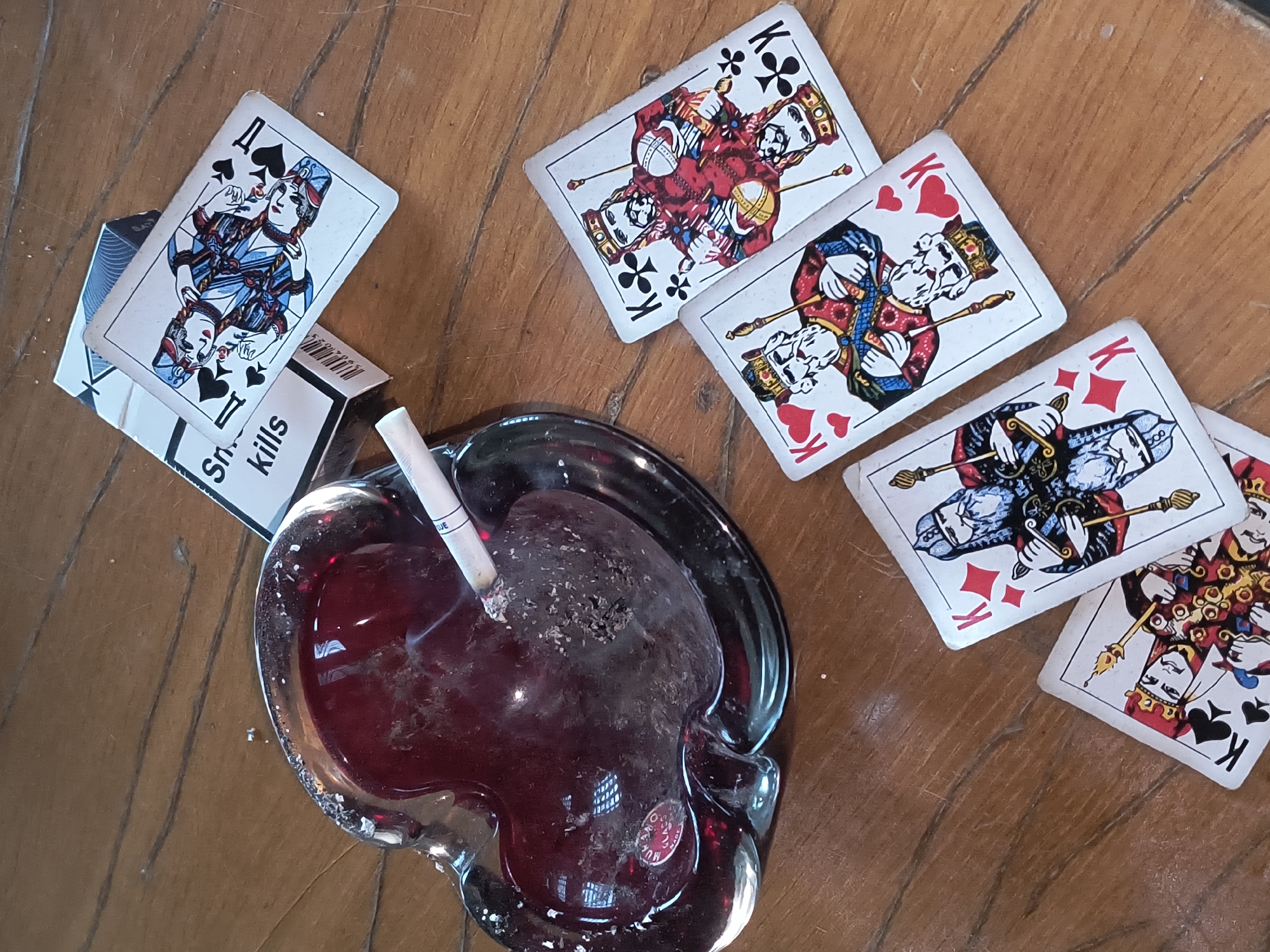 The image depicts a close-up overhead view of a wooden surface, likely a table, with various objects arranged on it. In the upper left is a heart-shaped, red glass ashtray containing a lit cigarette that is actively smoking. To the right of the ashtray, a pack of cigarettes lies on its side, with the brand name partially obscured, but the words "Sn." and "kills" are visible, suggesting a warning label. Several playing cards are scattered around the ashtray and cigarette pack. These include multiple King cards, identifiable by the "K" insignia and the depiction of kings, and at least one Queen card. The cards are spread out in a somewhat haphazard manner, with some overlapping. The lighting suggests an indoor setting, possibly with natural light coming from above or the side. There are no people visible in the image. The overall scene evokes a sense of casual gambling or a break for a cigarette. There is no specific indication in the image that places this scene in Rivne, Ukraine.
