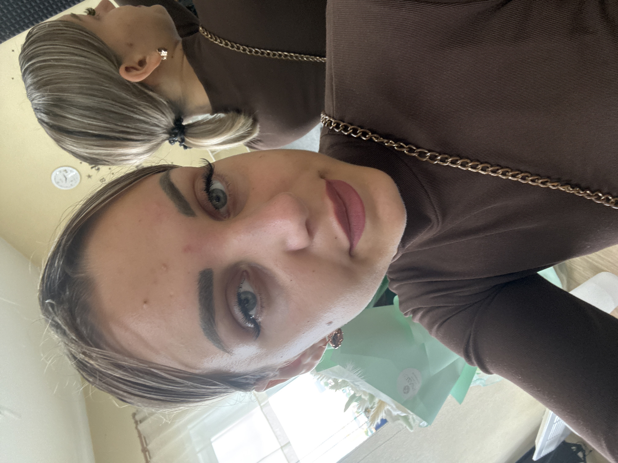 The image is a selfie taken by a young woman indoors. She is looking directly at the camera, with her face slightly angled. She has fair skin, light brown hair pulled back in a ponytail, and is wearing dark brown clothing with a gold chain strap visible. Her eyes are open and she has makeup on, including eyeliner and mascara. The background is somewhat blurred, but appears to be an indoor setting with a window and a mirror reflecting another part of her. There is also a bouquet of flowers wrapped in light green paper on the left side of the frame. The image does not provide any visual cues or context that would indicate its location in Yuzhno-Sakhalinsk, Russia.