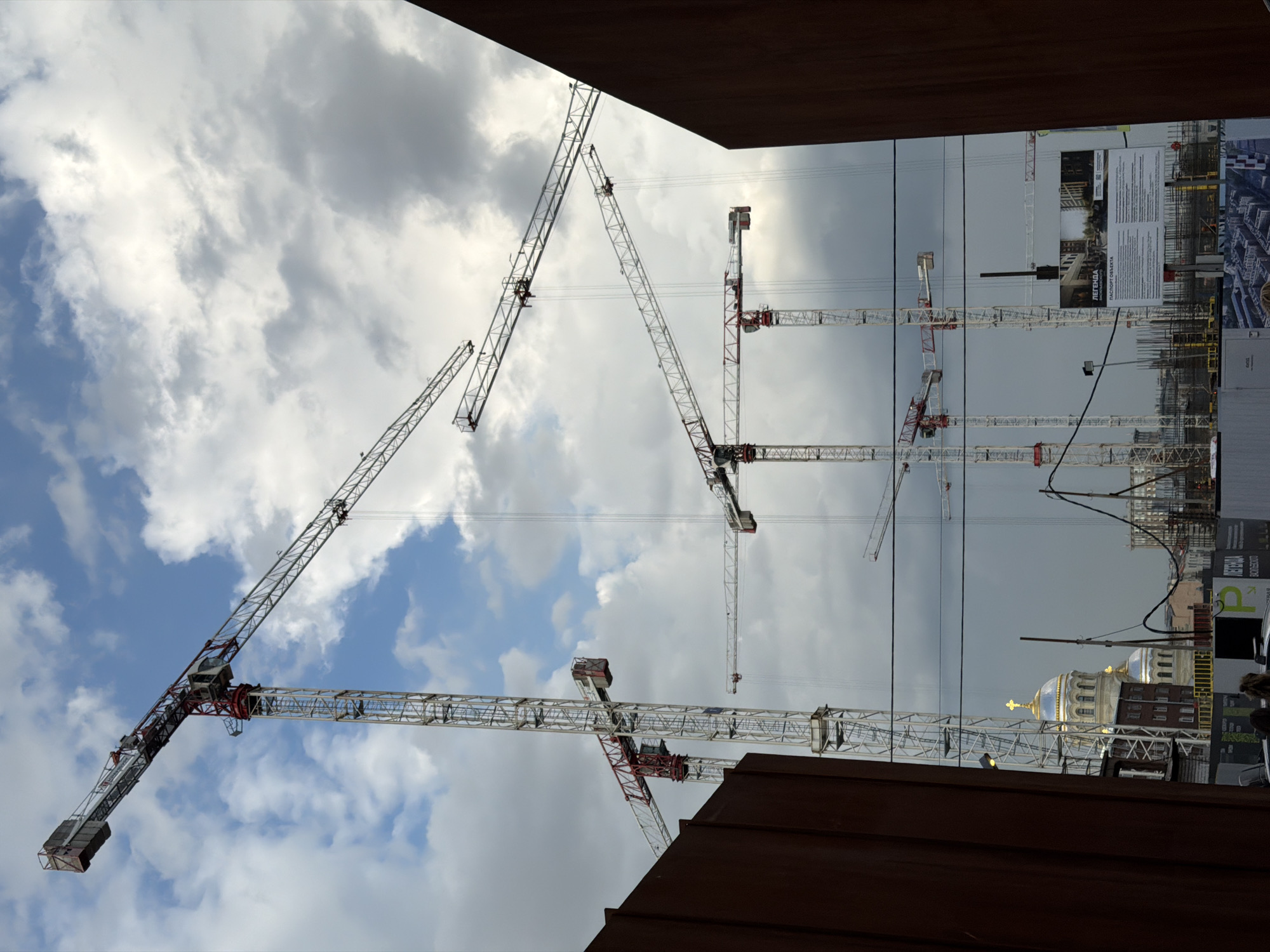 This media file captures a panoramic view of an active construction site in Saint Petersburg, Russia, framed by dark brown structures (possibly buildings or walls) at the top and bottom edges. The scene is dominated by a dense arrangement of at least seven white and red lattice tower cranes, with their booms and counterweights extending across the frame.
The sky above is a mix of bright white cumulus clouds against patches of blue sky on the left, transitioning to a more uniformly overcast gray on the right side of the image. Several thin wires or cables are visible traversing the sky horizontally.
In the mid-ground and background on the right, a distinctive historic building with two prominent golden domes, each topped with a cross, stands out, partially obscured by power lines and construction elements. This architectural style is characteristic of Russian Orthodox cathedrals. Below and around this, numerous partially constructed buildings, scaffolding, and other building materials indicate ongoing large-scale development.
A large construction information board, visible in the center-right, displays text in Cyrillic, including "ЛЕГЕНДА" (Legend) and "ПАСПОРТ ОБЪЕКТА" (Object Passport/Project Details), along with smaller, illegible descriptive text and architectural renderings. This signage reinforces the Russian location.
The image depicts a daytime scene, likely under variable cloudy conditions. The overall impression is one of dynamic urban growth, contrasting modern construction efforts with historical architectural heritage. No individuals are clearly visible.