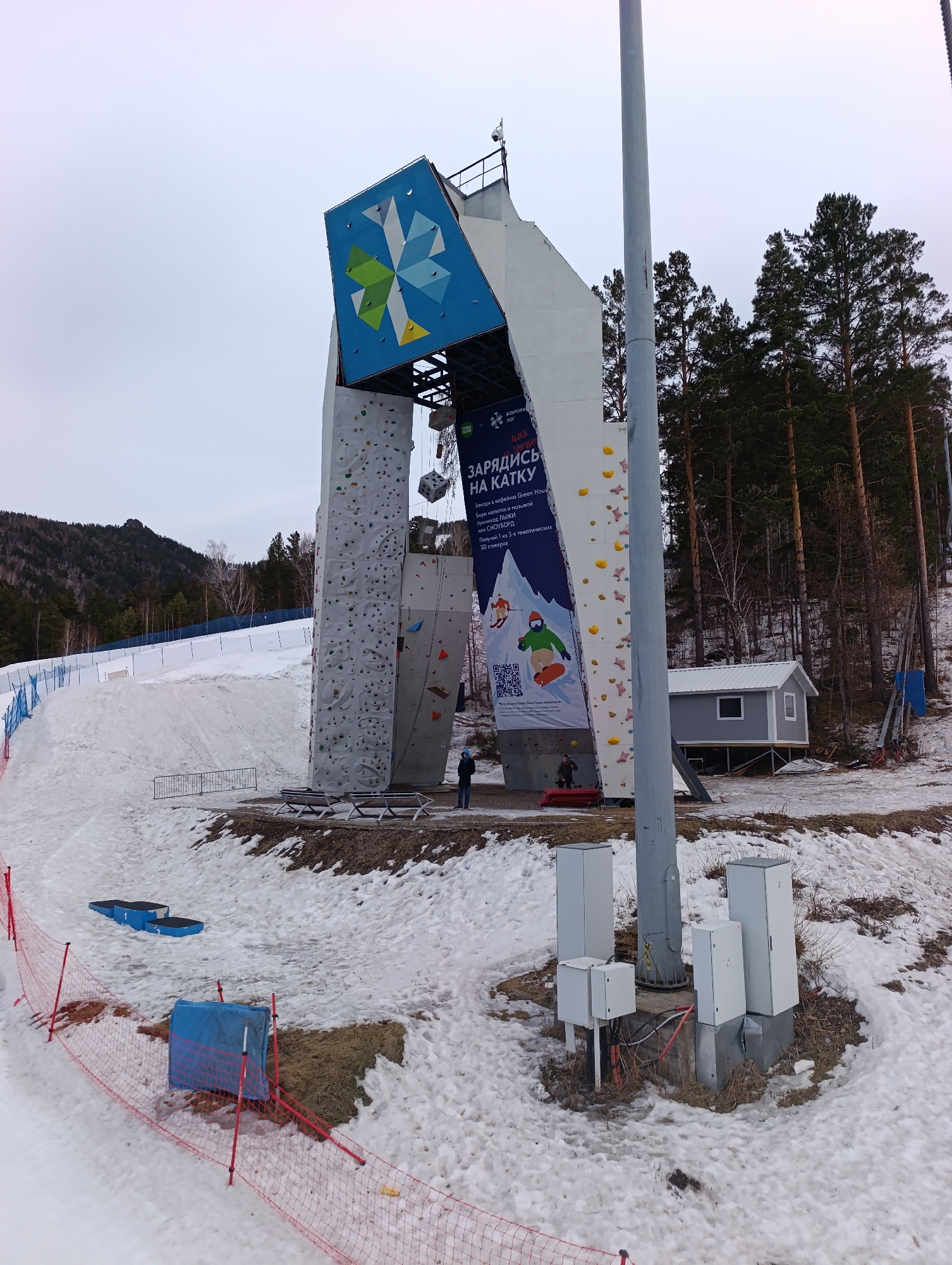 The image shows a climbing wall in what appears to be a recreational area in Krasnoyarsk, Russia. The structure is tall and features two distinct climbing surfaces. Above the climbing surfaces, there is a large, tilted sign with a blue and green geometric design. A banner hangs to the right of the climbing wall, advertising "Зарядись на Катку" (Charge on the ice rink), with a QR code and images of people skiing and snowboarding. Two people are present near the base of the wall. The surrounding environment is snowy, with patches of bare ground. A small building is located behind the climbing wall, and tall pine trees line the background. A red safety fence is visible in the foreground, along with some blue equipment or coverings. A tall utility pole stands near the climbing wall with several utility boxes at its base. In the distance, there are snow-covered hills and forested areas.