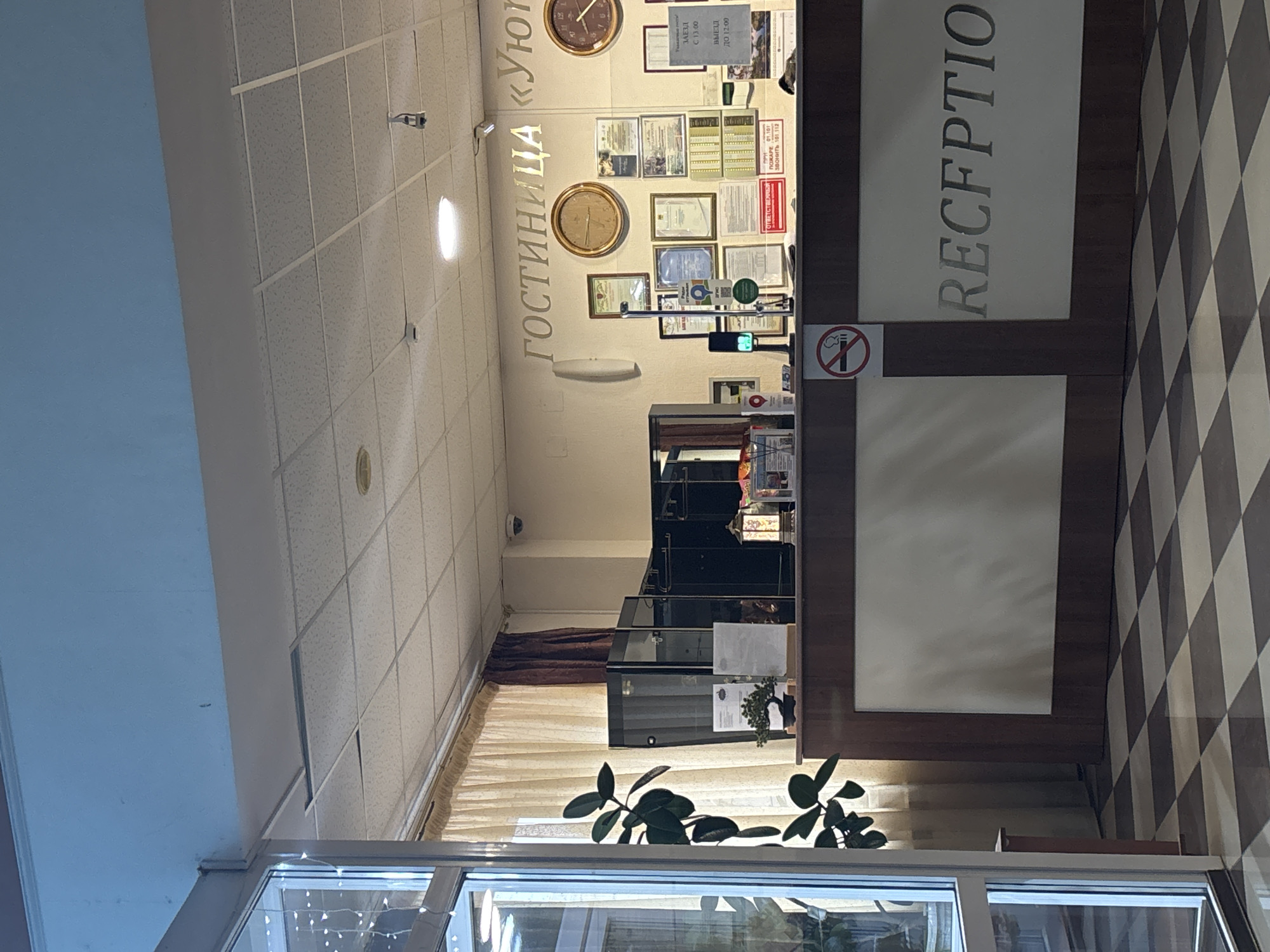 This interior shot depicts the reception area of the Uyut Hotel in Orenburg, Russia. The reception desk is on the right with the word "RECEPTION" vertically aligned on it. Above the desk are clocks and various framed documents. A non-smoking symbol is also visible. A plant sits in front of a window below the desk, and the floor has a checkered pattern. The background shows a curtained hallway leading further into the hotel.