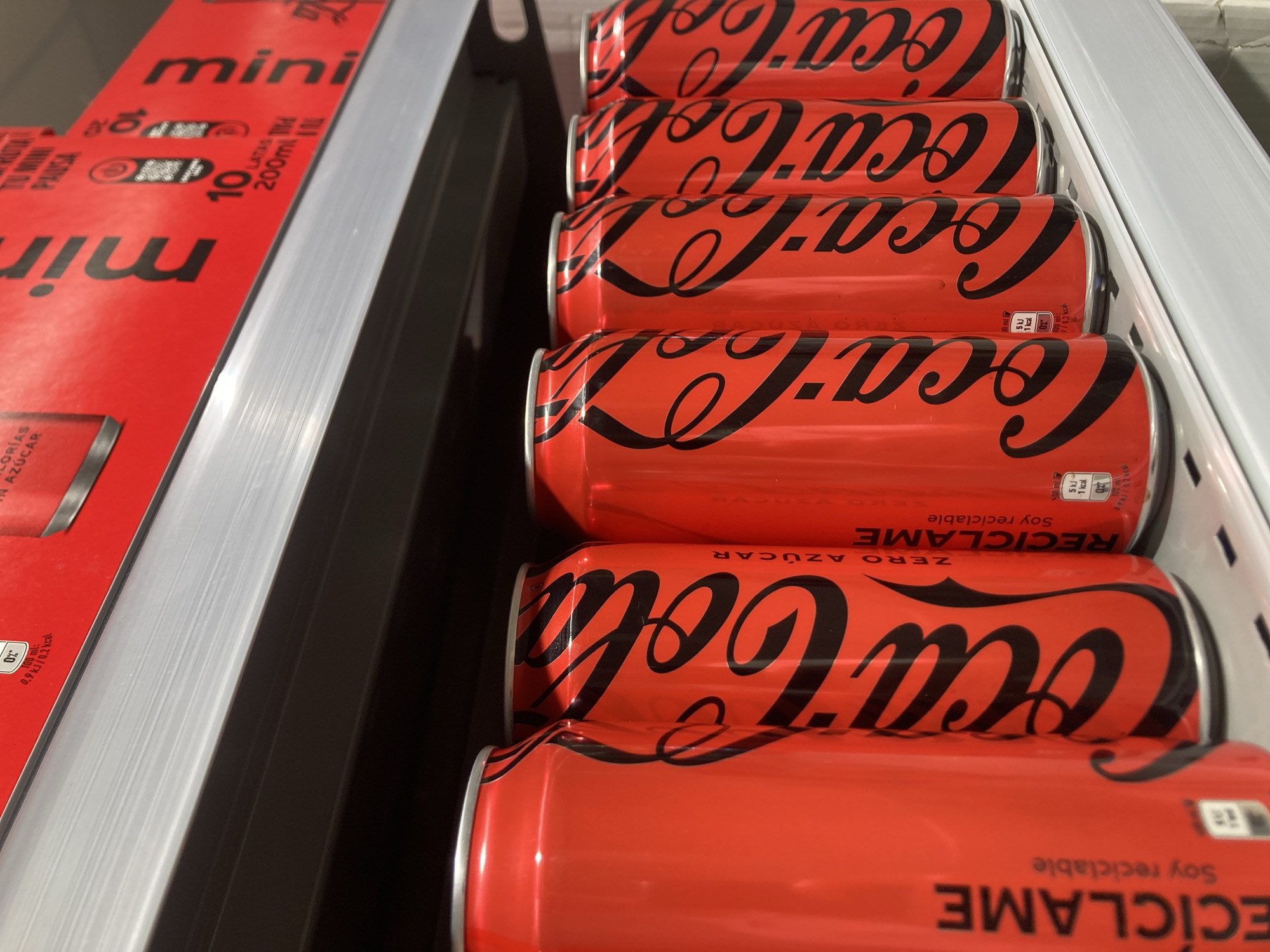 The image shows a close-up, eye-level view of several red Coca-Cola cans on a white shelf. The cans are arranged in a row, with the closest can on the left being partially visible. The Coca-Cola logo is prominently displayed on each can in black, cursive lettering. The cans appear to be the "Zero Sugar" variety, as indicated by text on some of them. The lighting suggests an indoor environment, possibly a supermarket or convenience store. The background is dark, with a slightly out-of-focus red box or packaging visible on a shelf above the cans. The overall impression is of a retail display of Coca-Cola products. The specific location within Barcelona, Spain cannot be determined from the image alone.