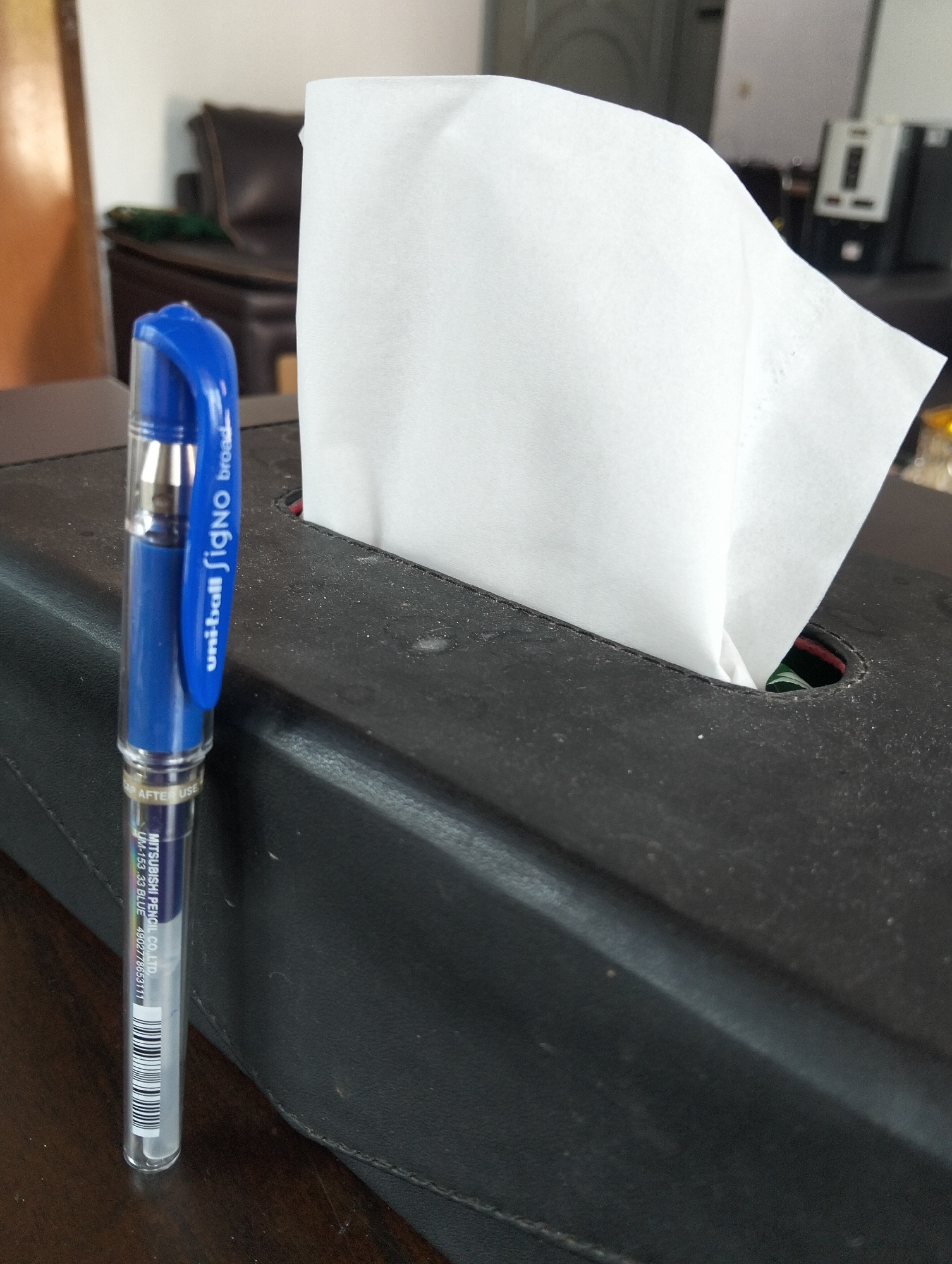 A blue and silver Uni-ball Signo broad pen stands upright in front of a black tissue box, with white tissues peeking out. The pen has a clear barrel revealing its inner workings and a blue cap. The tissue box appears to be made of faux leather and is dusty. In the background, a blurry image shows a dark brown leather armchair and a wooden door frame, suggesting an indoor setting. The scene is likely located in Julicotmeurak, Indonesia, though no specific landmarks are visible.
