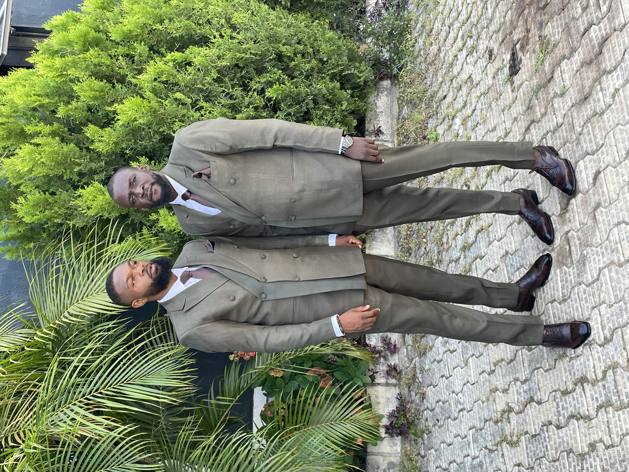 Two men, likely twins or closely related, are standing side-by-side in what appears to be an outdoor setting in Jos, Nigeria. Both men are wearing matching olive-green double-breasted suits, white dress shirts, and brown leather shoes. The man at the top of the frame has a watch on his left wrist. Both men have dark beards and short haircuts.
They are standing on a paved surface with interlocking bricks. Behind them, there is lush greenery consisting of various plants and trees, including a tall evergreen shrub and palm-like plants. The background suggests a well-maintained garden or park. The scene is well-lit, indicating it is daytime. The overall composition suggests a formal occasion or portrait setting.