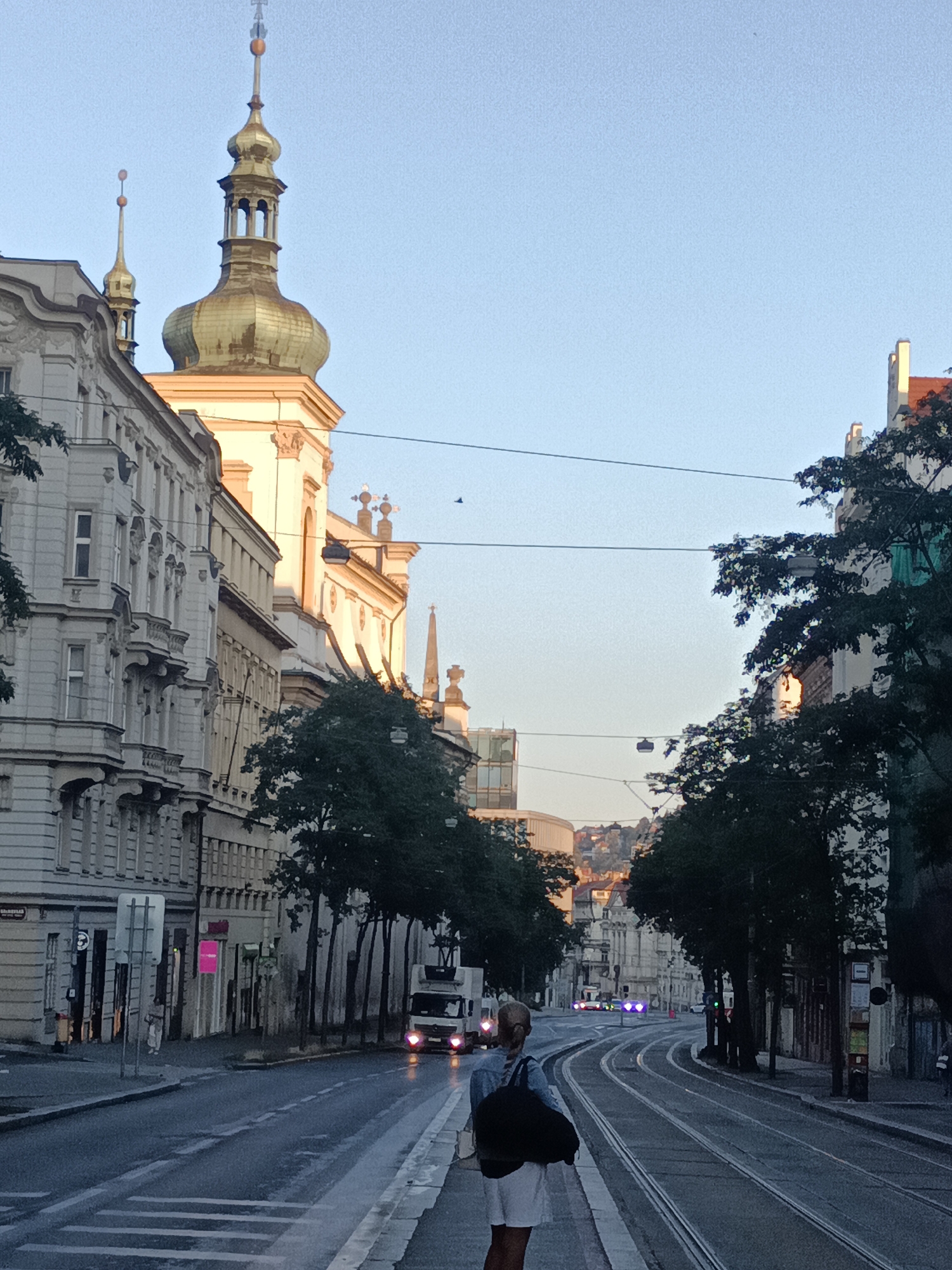 The image shows a street in Prague, Czechia, with tram tracks running down the middle. On the left, there are ornate buildings with classical architecture. On the right, there are trees and more buildings. A person is standing on the sidewalk, facing away from the camera, and carrying a black bag over their shoulder. A truck is visible in the distance, driving on the street. The sky is clear and blue, and the sun appears to be low in the sky, suggesting it is either early morning or late afternoon. The overall scene depicts a typical urban streetscape in Prague.