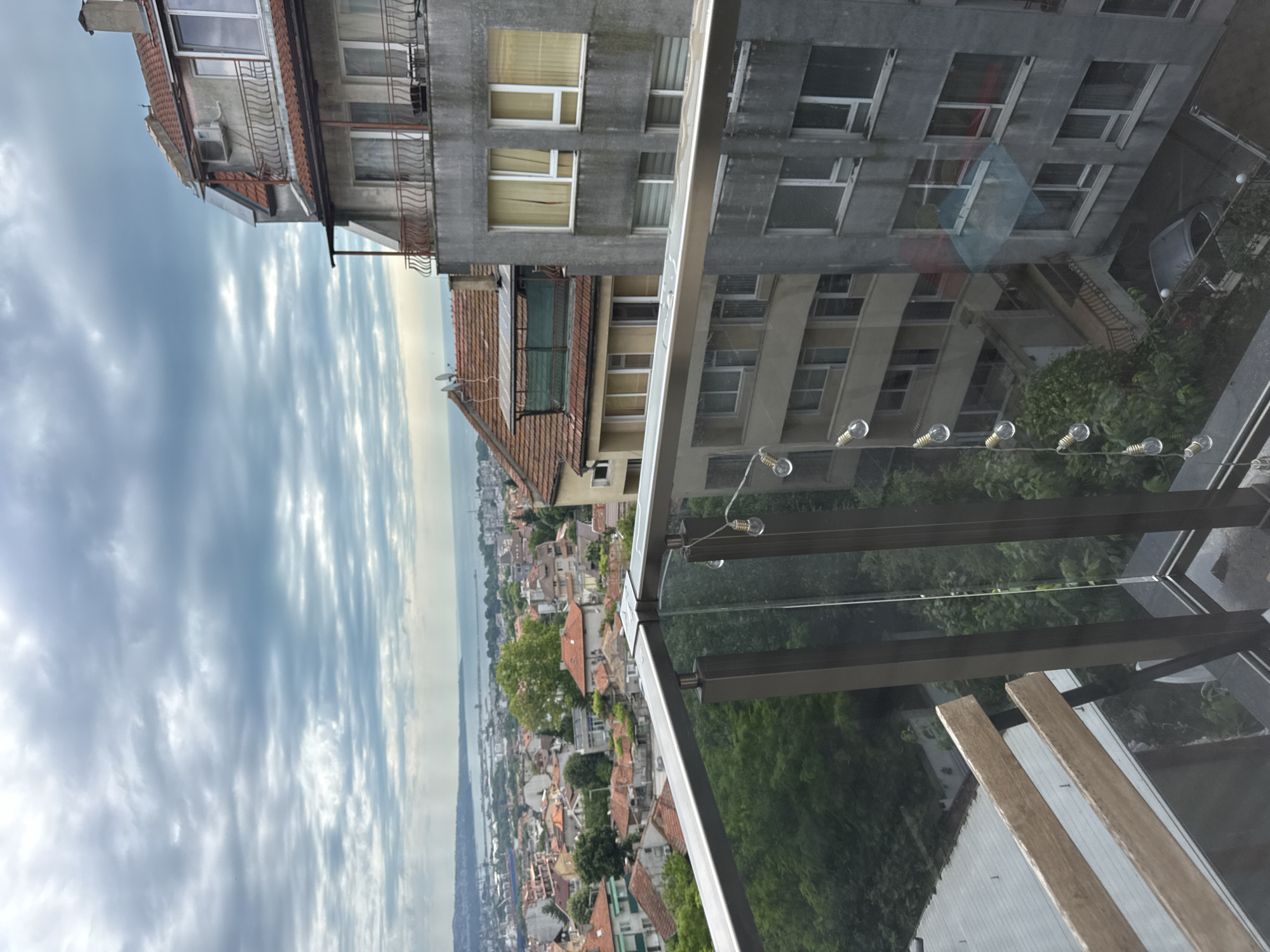 The image shows a view from a balcony overlooking a city with a body of water in the distance. The sky is cloudy, with a mix of grey and white clouds. In the foreground, there is a glass and metal railing with a string of lights attached. A wooden chair is partially visible to the left.
The city below is densely populated with buildings of varying heights and architectural styles. Many buildings have red-tiled roofs. There are trees interspersed throughout the urban landscape, indicating a moderate level of greenery. In the middle ground, a larger apartment building with a grey facade and numerous windows is prominent. Further in the distance, industrial structures and a large body of water, likely a sea or bay, can be seen. The overall impression is a coastal city with a mix of older and more modern architecture.