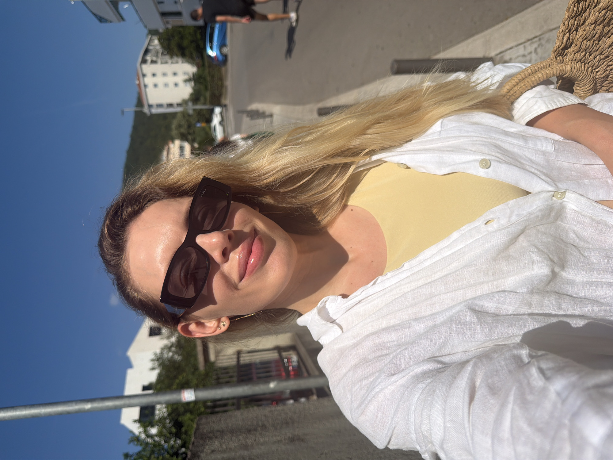 A young woman with long blonde hair and sunglasses takes a selfie outdoors on a sunny day. She is wearing a yellow top and an open white linen shirt. She has a straw bag slung over her shoulder. The background shows buildings, a street, and a blue car. The location is identified as Budva, Montenegro.