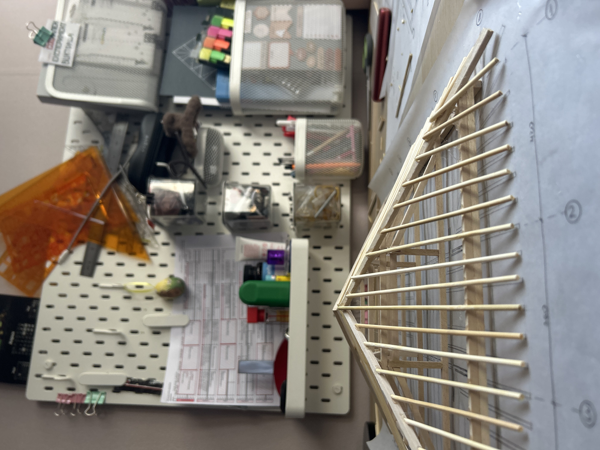 This image captures a busy and organized workspace, likely an architectural studio or craft area, with a pegboard organizing tools and a wooden model under construction. The overall perspective is a slightly downward, tilted view.
On the left and central part of the image, a white pegboard serves as a storage system. Several mesh containers are attached, holding various stationery items: colorful sticky notes (pink, orange, yellow, green), pens, and other small office supplies. A small, dark plush toy hangs from a hook. An orange, transparent drafting tool, possibly a set square or template, is visible near the top left. Lower down on the pegboard, sheets of paper with printed grids or tables are attached, alongside red and green binder clips, and a white container holding a red disk-shaped object and other supplies. Visible text, partially inverted and upside down, on a piece of paper attached by a binder clip reads "DICTIONARY".
To the right, a delicate wooden architectural model is being assembled. It appears to be a roof structure, with thin wooden sticks forming a framework of rafters. The model rests on or is positioned next to a sheet of paper featuring architectural plans or blueprints. On this paper, numbered circles like "①" and "②" are visible, along with the measurement "420".
The scene suggests an ongoing creative or design project, characterized by meticulous organization of materials on the pegboard and the detailed construction of the model. The lighting is consistent with an indoor setting. No specific location cues for Sofia are visible within the image.