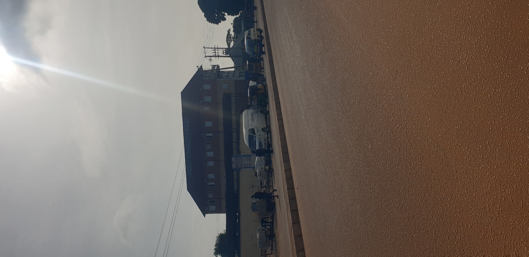 This street scene, captured in what is likely Sabon Tasha, Nigeria, presents a daytime view under an overcast sky, with a strong sun glare originating from the upper left, casting a bright streak across the clouds. The foreground and midground on the right are dominated by a wide, reddish-brown, textured road, which appears to be unpaved or made of laterite.
Along the left side of the road, a multi-story building with a dark roof and numerous windows forms a significant part of the background. In front of this building, various vehicles are visible. A white van, clearly marked "COMMAND COLLEGE" on its side, is prominent in the mid-ground. Nearby, a signpost displays "DULLOMA PETROLEUM" with a list of what appears to be fuel prices for "PMS," "AGO," and "DPK," followed by numeric values such as 780, 110, 490, 100, and 570.
Several auto-rickshaws, locally known as Keke Napeps, are parked or queuing along the street behind the white van. Numerous people are present, standing and sitting near the vehicles and beside what seem to be informal market stalls or street vendor setups, characterized by large sacks or bundles of goods. Electrical poles and overhead wires crisscross against the sky and buildings. Trees are scattered in the background, adding some greenery to the urban landscape. The overall impression is a bustling street in a commercial or community area.