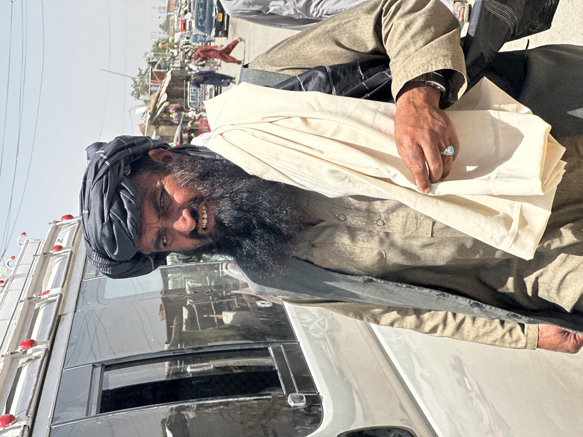 In Kanozai, Pakistan, a man with a dark beard and a black turban walks on a sunny street. He is wearing a brown tunic and a grey vest over it. He has a light-colored cloth draped over his shoulder and arm. His left hand, adorned with a turquoise ring, rests on the cloth. In the background, a silver van is parked, and a busy street scene unfolds with vehicles, market stalls, and other pedestrians under a clear sky.