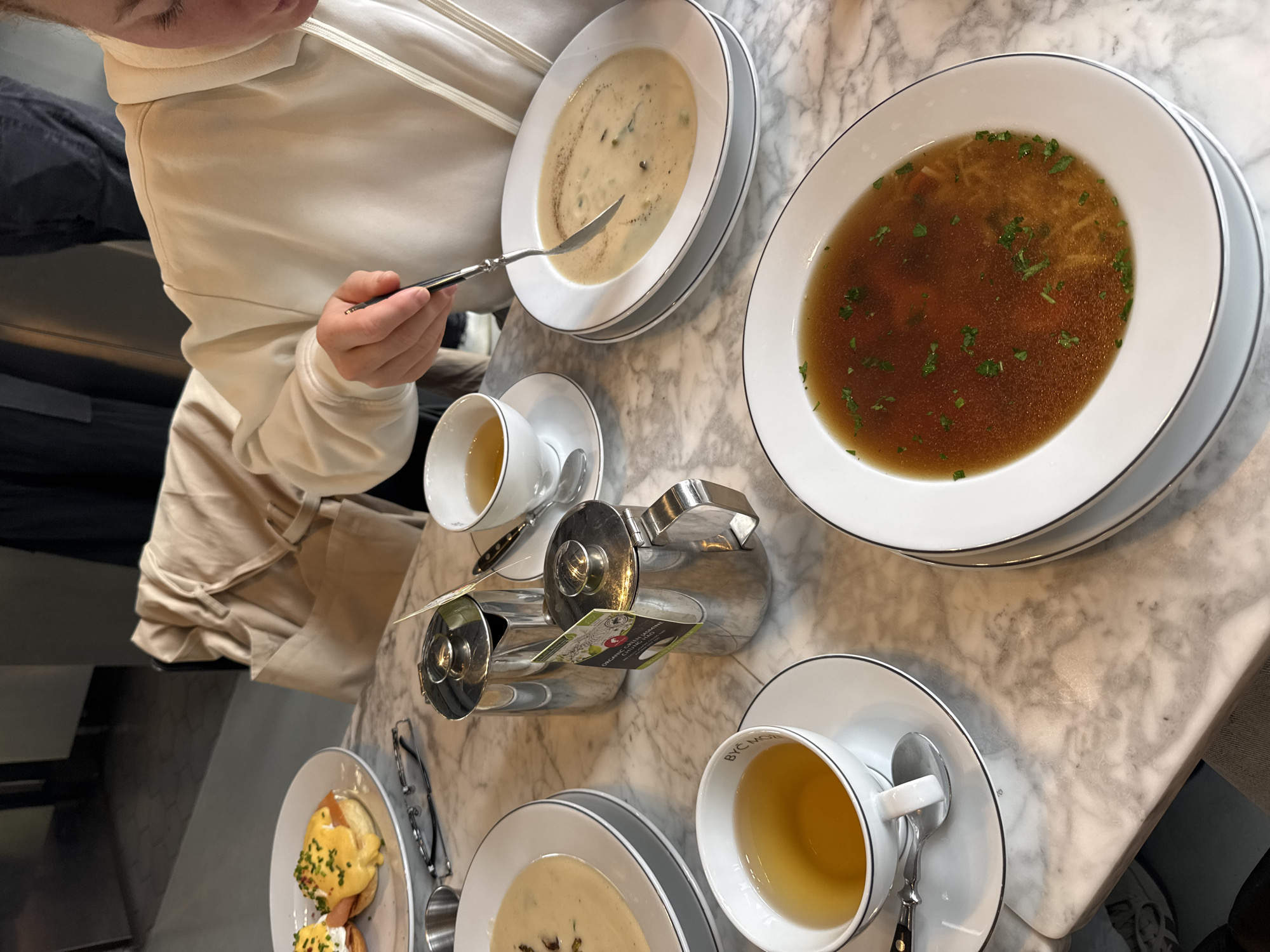 A photograph shows a dining table at a restaurant, possibly in Warsaw, Poland. A person wearing a white jacket and a watch on their left wrist is seated at the table and is eating a bowl of soup with a silver spoon. The table is made of light gray marble. Other food items include two plates of soup (one light-colored creamy soup and one brown soup), and a plate of Eggs Benedict with sauce. There are also two cups of tea and a teapot on the table. The plates and cups are white with a black rim. The lighting appears to be natural. The overall impression is one of an elegant dining experience.