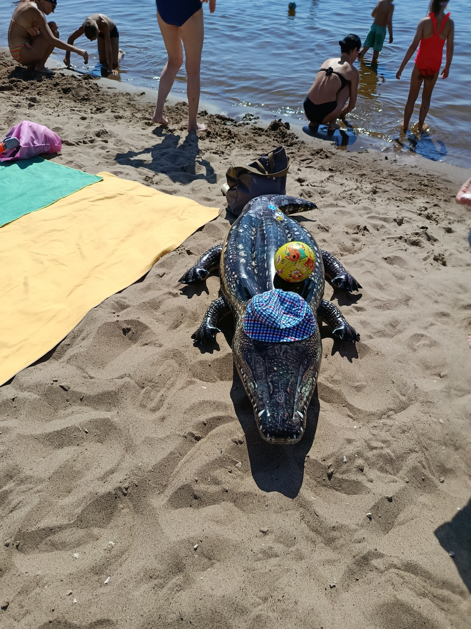 An inflatable alligator float lies on a sandy beach next to a yellow and a green towel. The alligator is dark colored with white speckles and has a blue and white checkered hat on its head. A colorful ball sits on its back. In the background, several people are near the water, with one person sitting on the sand near a pink bag. The water appears calm and the sky is clear. The scene is set outdoors on a sunny day.