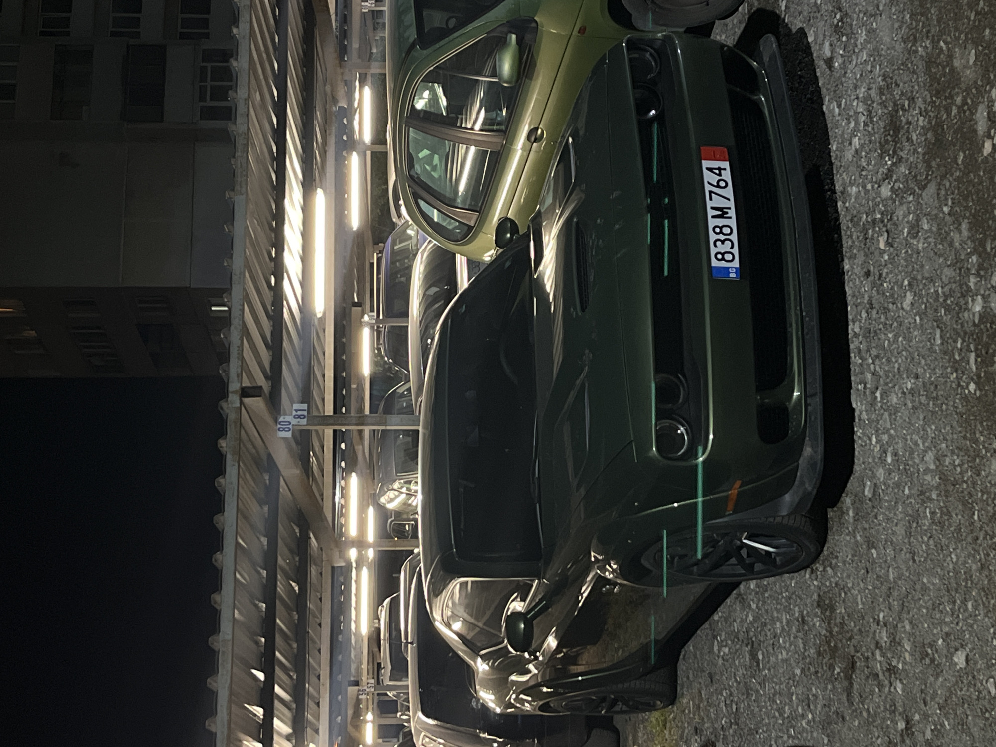 The image depicts an indoor parking lot at night. A dark green Dodge Challenger with Bulgarian license plate "838 M764" is parked in the foreground. Other cars are parked nearby, including a green hatchback partially visible above the Challenger. Overhead, the parking structure features bright fluorescent lights illuminating the scene, along with parking spot numbers like "80-81". A building is visible in the background to the left. Based on the license plate, the location is likely in Bulgaria. The specific city, Sofia, is specified in the prompt. The scene is static and focuses on the parked vehicles.