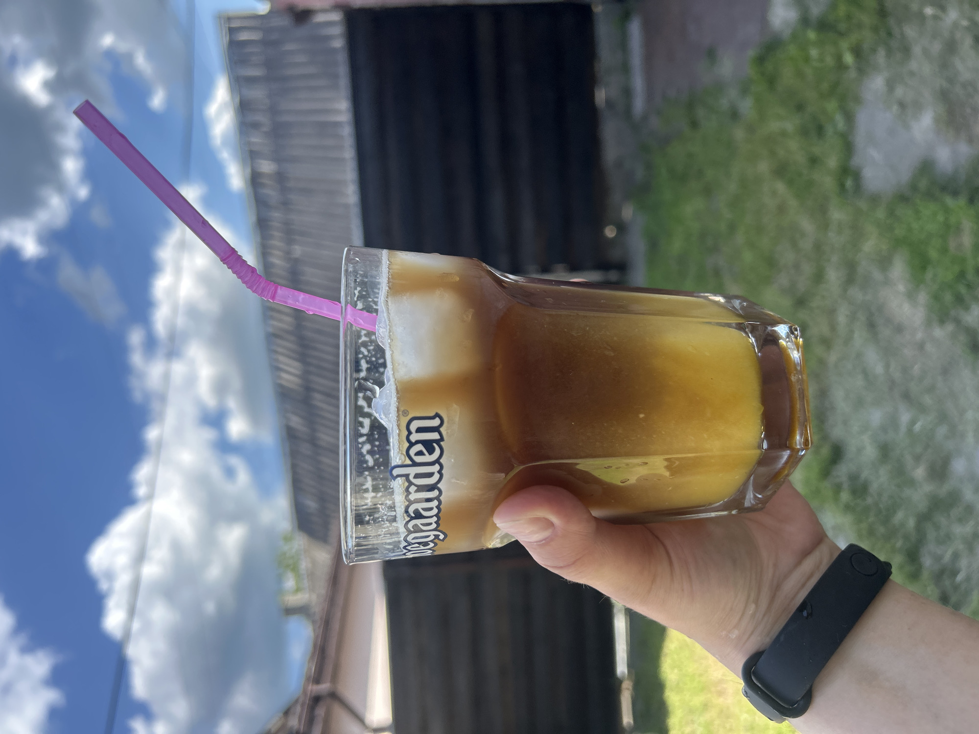 A hand wearing a black fitness tracker holds a glass of iced beverage with a pink straw. The glass has "Hoegaarden" printed on it. The beverage appears to be layered with a darker liquid on top and a lighter, possibly orange, liquid at the bottom, with ice cubes mixed in. The background shows a wooden fence, a slanted roof, and a partly cloudy blue sky. The scene is outdoors, likely in a backyard, with green grass in the foreground. The location is Bila Tserkva, Ukraine.