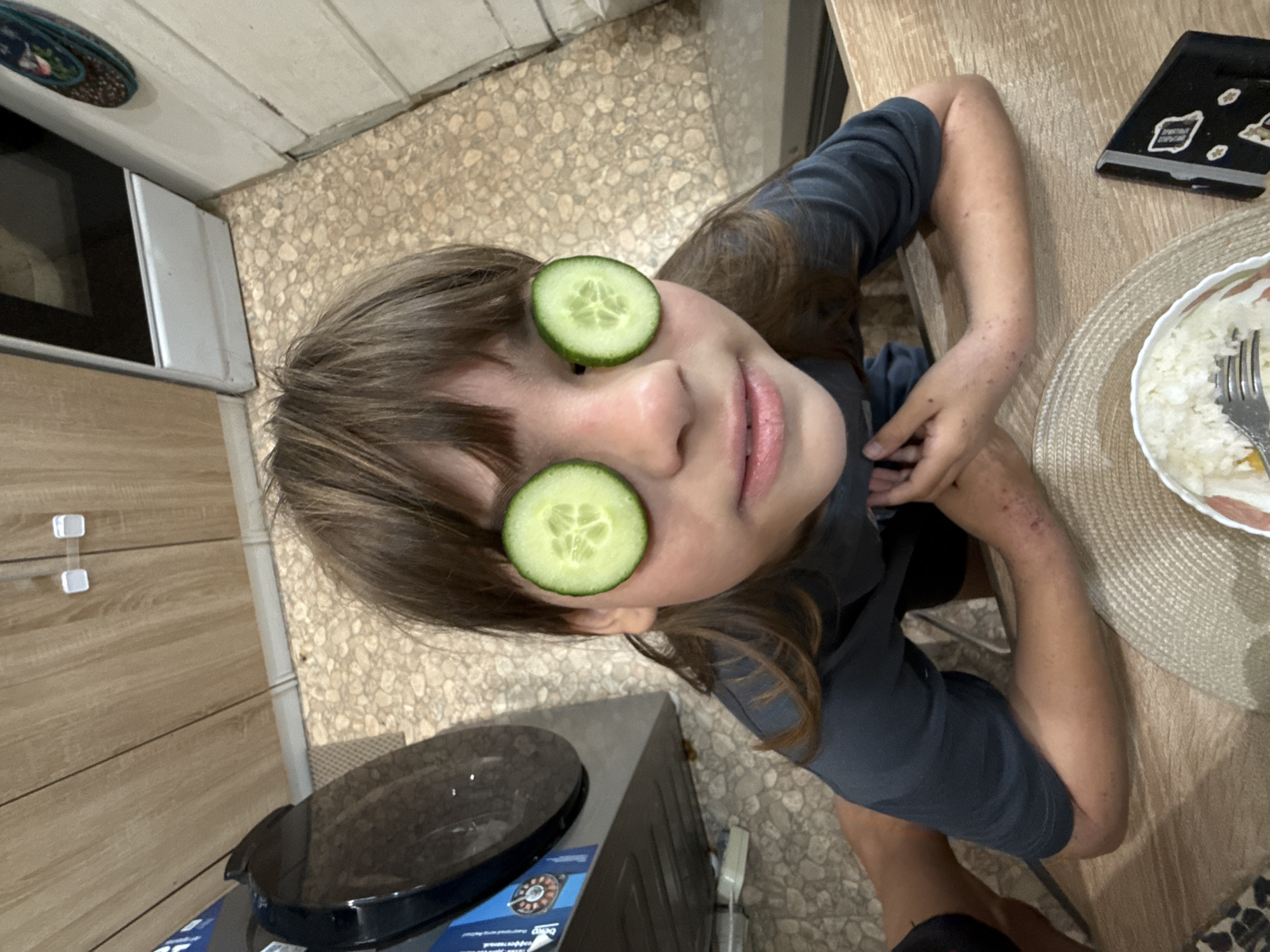 A young, light-skinned person with brown hair sits at a wooden table. The person has a slice of cucumber over each eye. A plate of food and a cell phone are on the table. The person is wearing a dark grey shirt. A refrigerator and floor tile are visible in the background. No information about the location of the scene being in Rusanovo, Russia, is given.