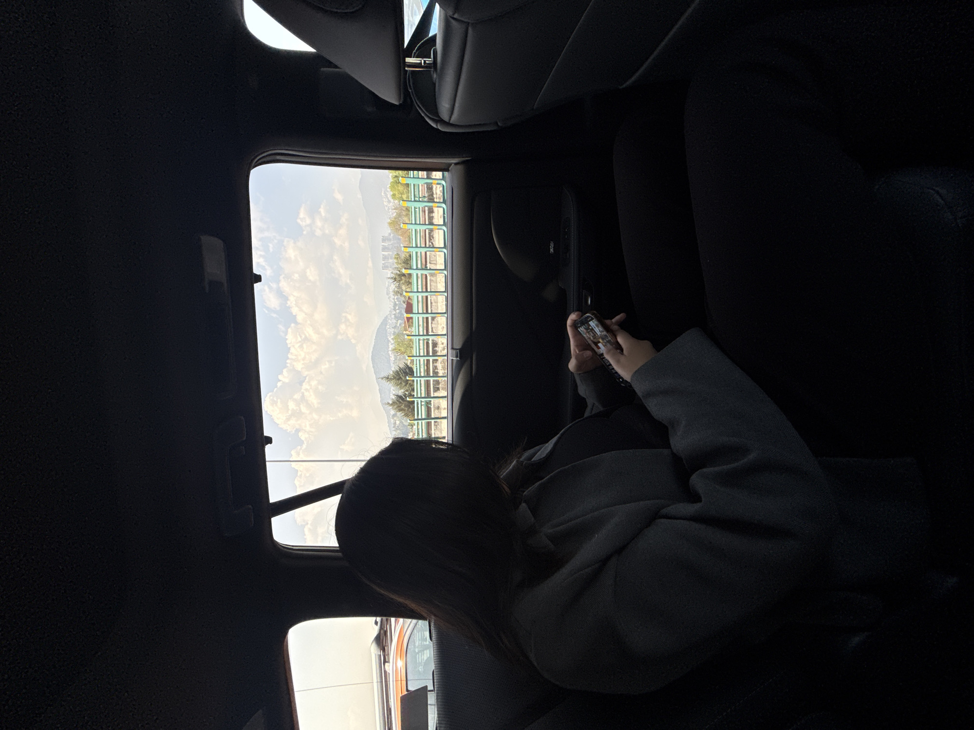 The image, rotated 90 degrees clockwise, captures a moment inside a vehicle, likely in Mexico City, Mexico. The primary subject is a person, appearing to be female with dark hair, seated in what looks like the front passenger seat or a rear seat. She is wearing a dark grey jacket or sweatshirt over dark pants. Her head is bowed, and she is engrossed in her smartphone, which she holds with both hands. The screen of the phone displays an image, possibly a selfie or another person, though details are indistinct.
The setting is the interior of a car, characterized by dark upholstery on the seats and ceiling. A side window offers a view of the outside. Beyond the car, the scene depicts a bright, sunny daytime with a clear sky adorned with fluffy white clouds. A prominent feature in the mid-ground is a series of green and white railings or barriers, possibly part of a park, public space, or elevated structure. In the distance, green, rolling hills or mountains rise, with some urban buildings visible between them and the immediate foreground. The overall impression is one of travel or transit, with the person occupied by their device while the world passes by outside. No discernible text is legible other than on the smartphone screen, which is too small to read.