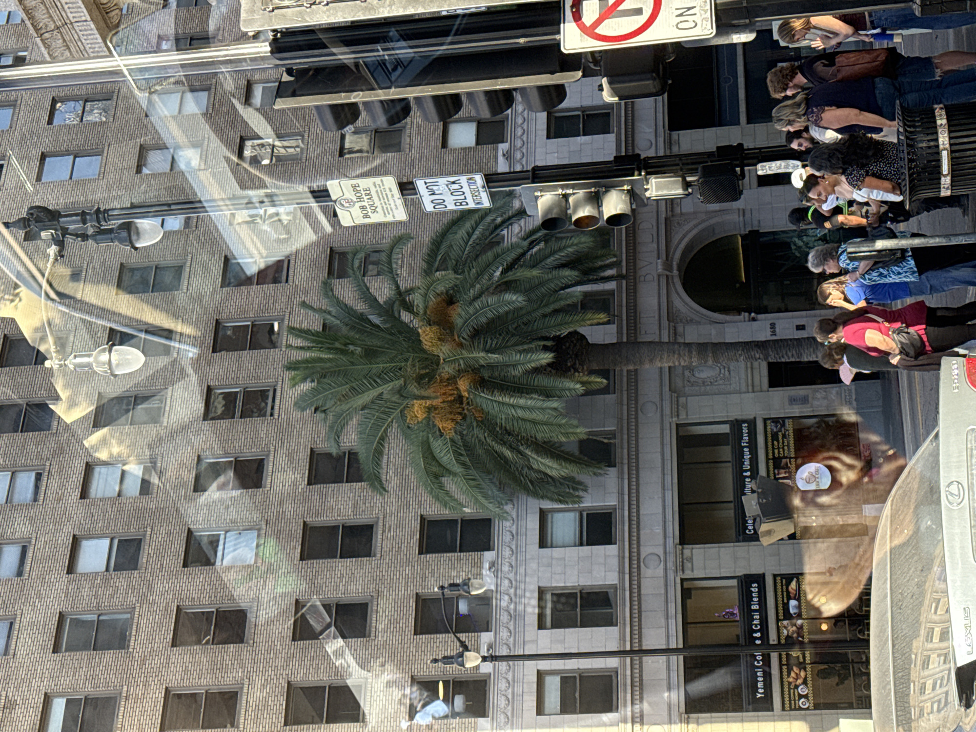 This image depicts a street scene in the city of Los Angeles, California. The focus is on a tall building constructed of beige brick, with numerous windows. In front of the building, a palm tree stands prominently. A street lamp and a traffic light are visible, along with various street signs. One sign reads "BOB HOPE SQUARE" and another displays "DO NOT BLOCK INTERSECTION." A group of people are standing on the sidewalk, some looking in different directions, and one person is holding a pink fan. Several cars are present, including a silver Lexus in the foreground. The overall scene suggests an urban environment with typical city elements.