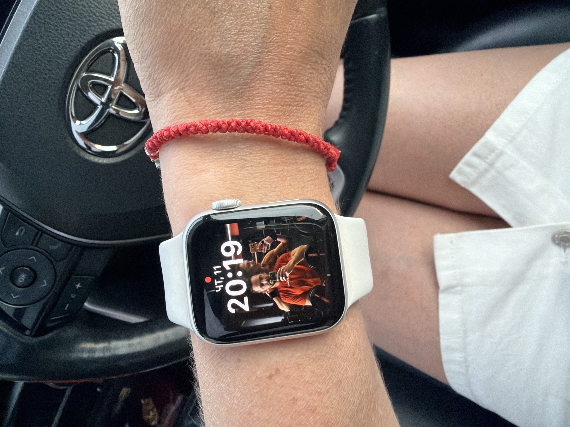 The image shows a close-up of a person's wrist wearing a white Apple Watch. The watch face displays the time as 20:19 and shows a mirror selfie of a man in an orange shirt. A red braided bracelet is also visible on the wrist. In the background, the steering wheel of a car with a Toyota logo is visible. The context of Birobidzhan, Russia is not determinable from the image.