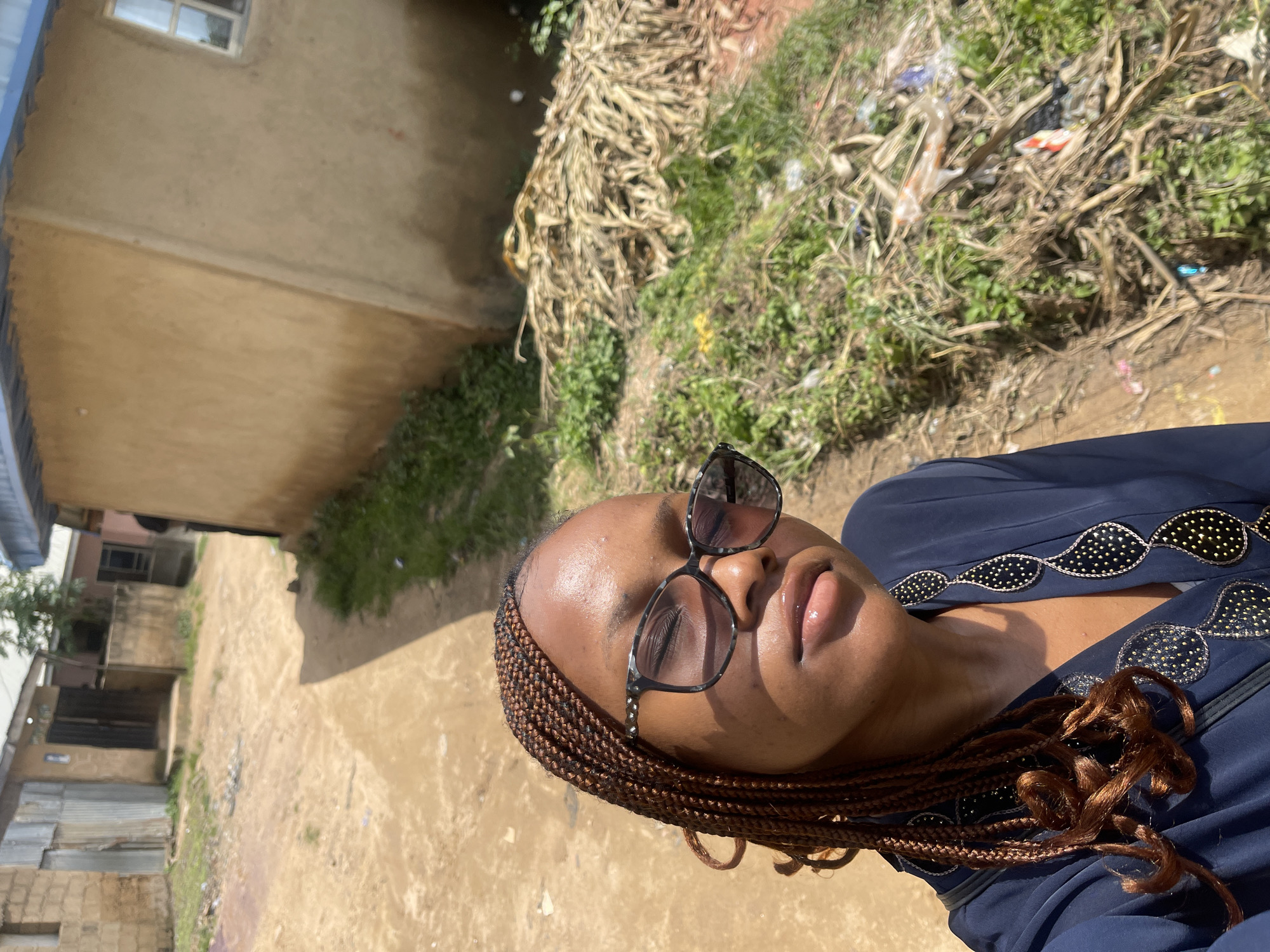 The media file shows a close-up, high-angle shot of a young Black woman wearing sunglasses and a dark blue dress. She has braided hair. Her face is angled upwards, with her eyes closed and lips slightly parted, suggesting a moment of relaxation or contemplation. The background is outdoors, featuring a dirt path, dry vegetation, some green plants, and evidence of scattered trash. To the left of the frame, a portion of a building with a window is visible, along with other structures that appear to be houses or small shops, suggesting an urban or semi-urban environment. The lighting suggests it is daytime, with bright sunlight casting shadows. The overall scene gives a sense of a casual, possibly warm day. No visible text is present in the image. The location context provided is Jos, Nigeria.