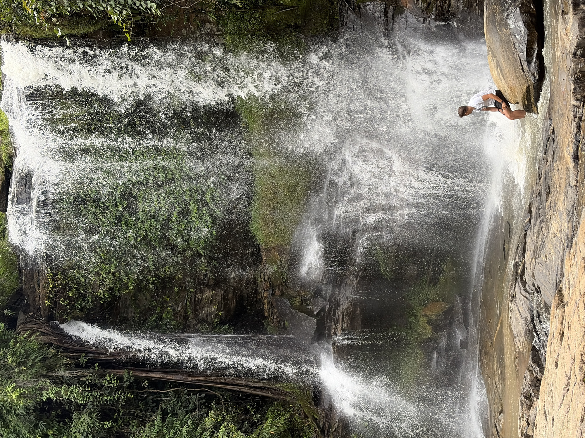 A person is sitting on a large rock at the base of a waterfall. The person is wearing a white t-shirt and black shorts. Their legs are submerged in the water. The waterfall is surrounded by lush green foliage and moss-covered rocks. The water is cascading down a rocky cliff face, creating a misty spray. The ground in the foreground is composed of large, flat rocks. The scene appears to be in a natural, outdoor environment, possibly a park or a jungle. This location is identified as being in the city of Ban Huai Takhrai, Thailand.