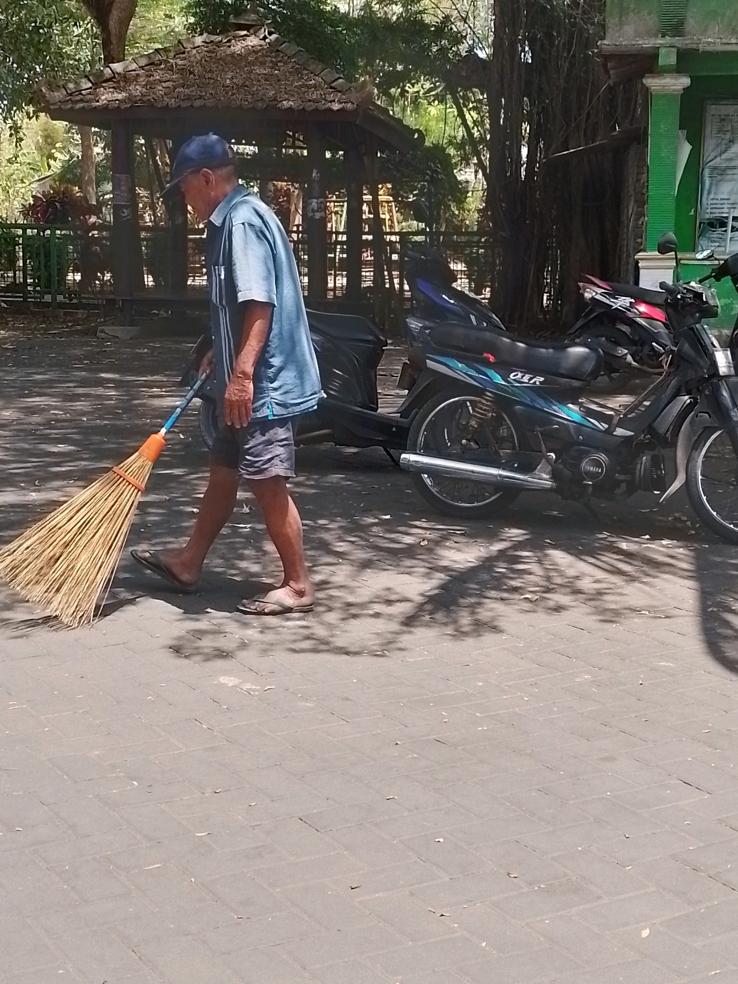 An elderly Indonesian man wearing a blue cap and a short-sleeved blue shirt is sweeping the ground with a broom. He is wearing grey shorts and flip-flops. The man is walking from left to right across a paved area. Behind him, to the right, are several parked motorcycles, including a black Yamaha. In the background, there is a thatched-roof pavilion and lush green foliage. The scene appears to be outdoors, possibly in a public area like a park or a street in the city of Wonosari, Indonesia. The ground is paved with interlocking bricks, and shadows from trees are cast across the area.