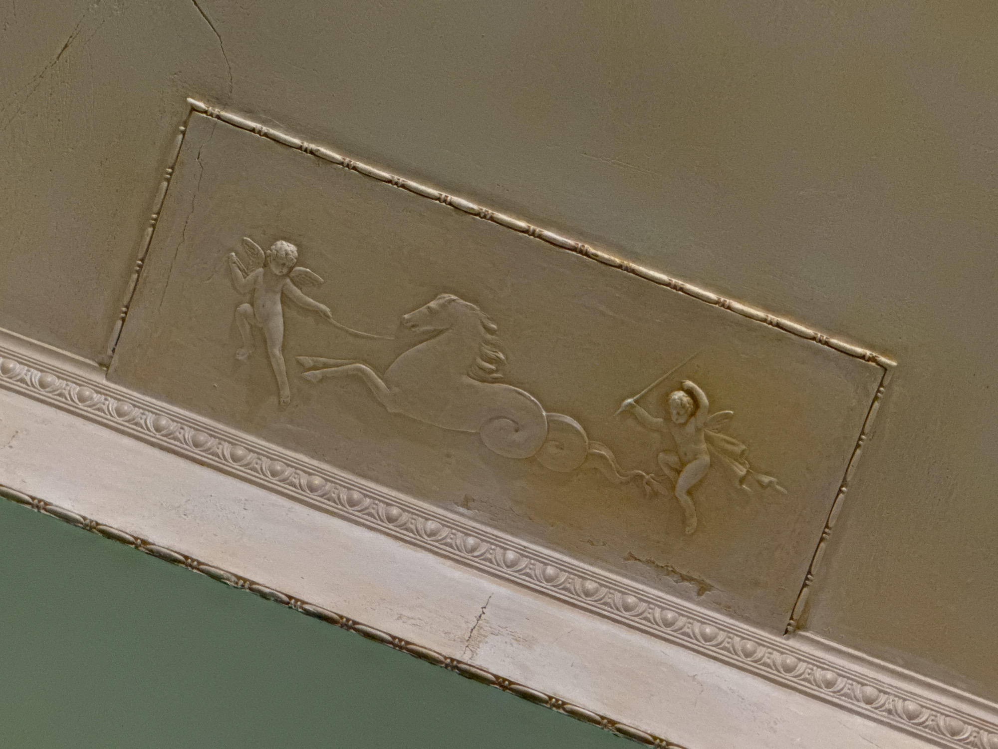 The image shows a close-up view of an ornate plaster relief on a ceiling. The relief depicts two cherubic figures and a creature resembling a sea horse. The figures and creature are rendered in profile, with the cherubic figures appearing to interact with the sea horse. The relief is set within a rectangular recessed panel, bordered by decorative molding. The ceiling itself is painted a muted tan color, and there are visible cracks in the plaster. The surrounding area of the ceiling is painted a light green. The style of the relief suggests it is from a historical building, likely a palace or grand residence.
This image was captured on the ceiling of the Grand Ballroom in the Yusupov Palace on the Moika River embankment in Saint Petersburg, Russia. The Yusupov Palace, a former aristocratic residence, is renowned for its lavish interiors and historical significance, including its association with the assassination of Grigori Rasputin. The Grand Ballroom, in particular, is known for its opulent decoration, including elaborate stucco work and chandeliers.