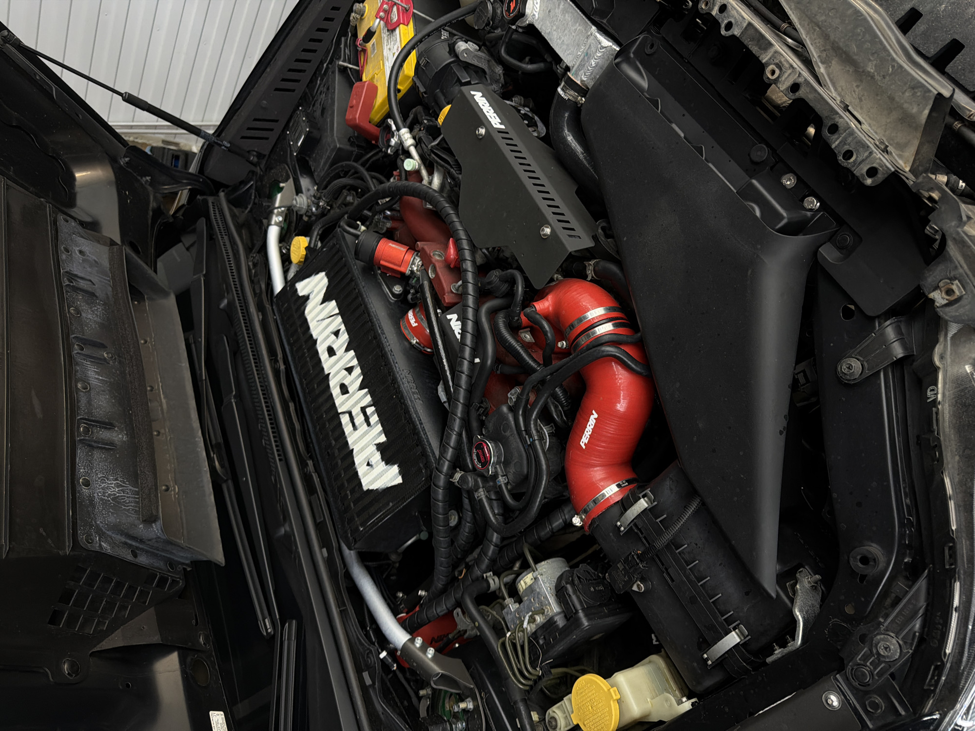 This media file presents a detailed view of a highly customized and meticulously clean car engine bay, seen from an elevated perspective with the hood open. The engine bay features a striking black and red color scheme, indicating significant aftermarket modifications.
The central and most prominent object is a large, black top-mount intercooler, clearly labeled with the white "PERRIN" logo. Connected to various components are bright red silicone intake pipes and hoses, also branded "PERRIN," which add a vibrant contrast to the otherwise dark engine components. A black engine cover or heat shield, also bearing the "PERRIN" logo, is visible further back.
In the upper-left, a yellow car battery with red positive and negative terminals is visible. Near the battery, a fluid cap with the "MOROSO" brand name is identifiable. A red filler cap, marked with the "STI" (Subaru Tecnica International) logo, suggests the vehicle is likely a Subaru performance model. In the lower-left, a silver strut brace is partially visible, indicating further performance enhancements. Various other components, including black plastic air intake boxes, black hoses, and electrical wiring, are neatly arranged throughout the bay. A yellow cap on a translucent fluid reservoir can be seen in the lower-right.
The setting appears to be indoors, likely a garage or workshop, as suggested by the bright, even lighting and the white, paneled wall visible in the background through the open hood. The overall condition of the engine bay is immaculate, suggesting recent work, maintenance, or preparation for display. The image captures the vehicle's engine in Krasnoyarsk, Russia, highlighting its customized and performance-oriented nature.