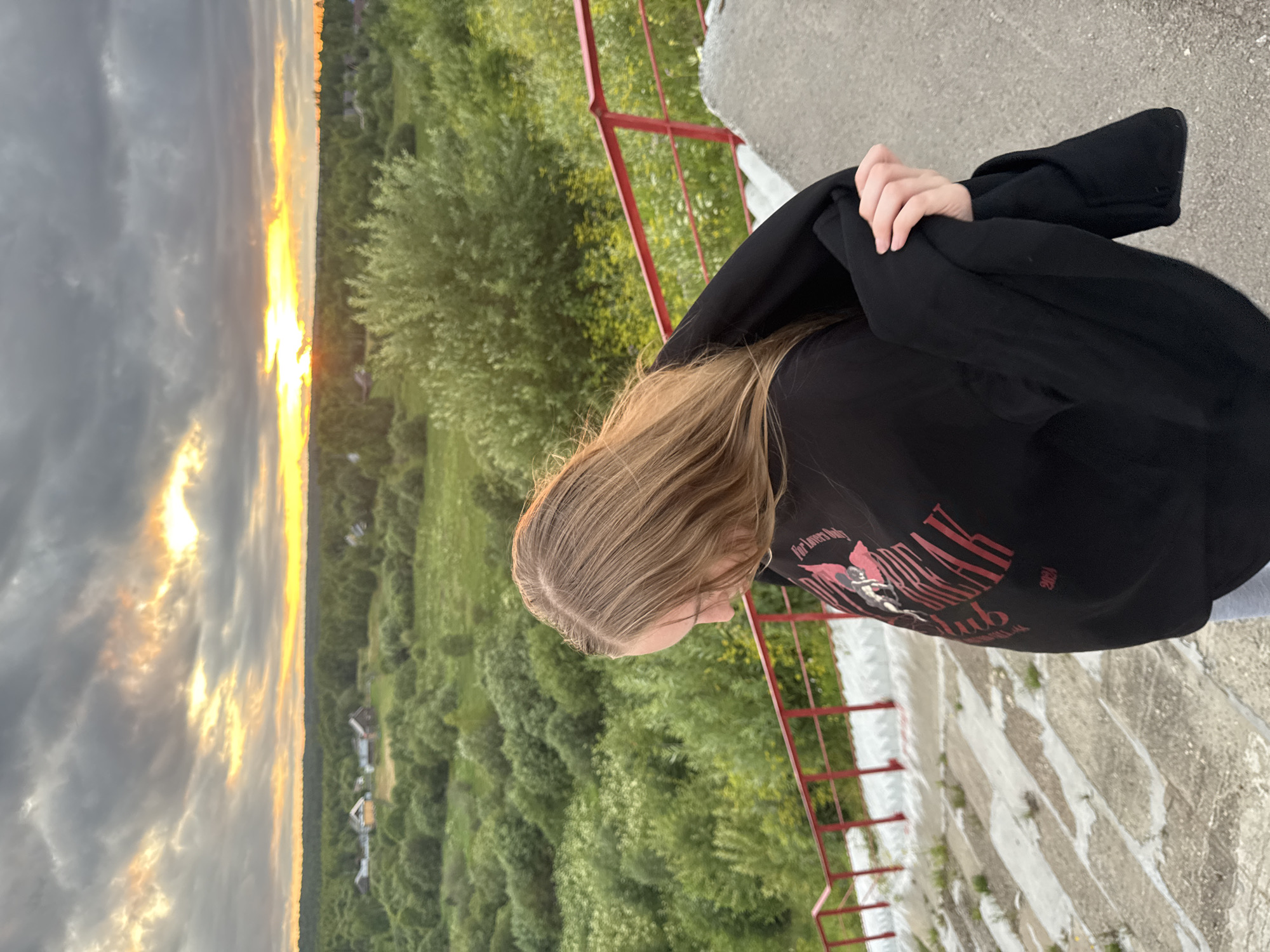 A young woman with blonde hair, seen from behind and slightly to the side, stands on a concrete staircase with a red railing. She is wearing a black sweatshirt with red lettering and holding a black garment. She is looking down and to her left. The staircase leads down to a landscape of green fields and trees under a cloudy sky with the sun setting and casting a warm glow. In the distance, there are small houses, suggesting a rural or semi-rural location in the city of Ryabinino, Russia. The overall scene depicts a serene moment at dusk in a natural setting.