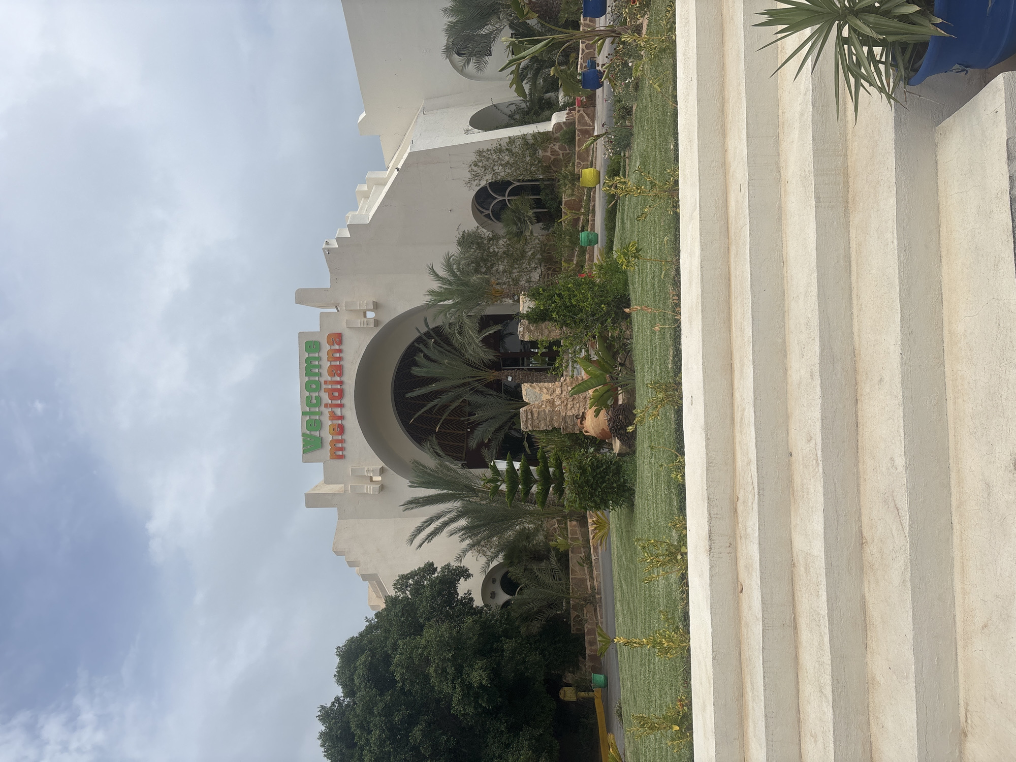 The image shows the exterior of a building with large arched entrances, likely a hotel or resort, located in Taguermess, Tunisia. The building is white with decorative stepped architectural elements. Above the central entrance, the words "Welcome Meridiana" are displayed in colorful lettering. The entrance area is surrounded by lush green grass, palm trees, and various plants. In the foreground, there are wide, light-colored concrete steps leading up towards the building. The sky is overcast with a few patches of blue visible. There are no people visible in the image. Various potted plants and some decorative elements, such as a large clay amphora, are placed around the entrance and landscaping.