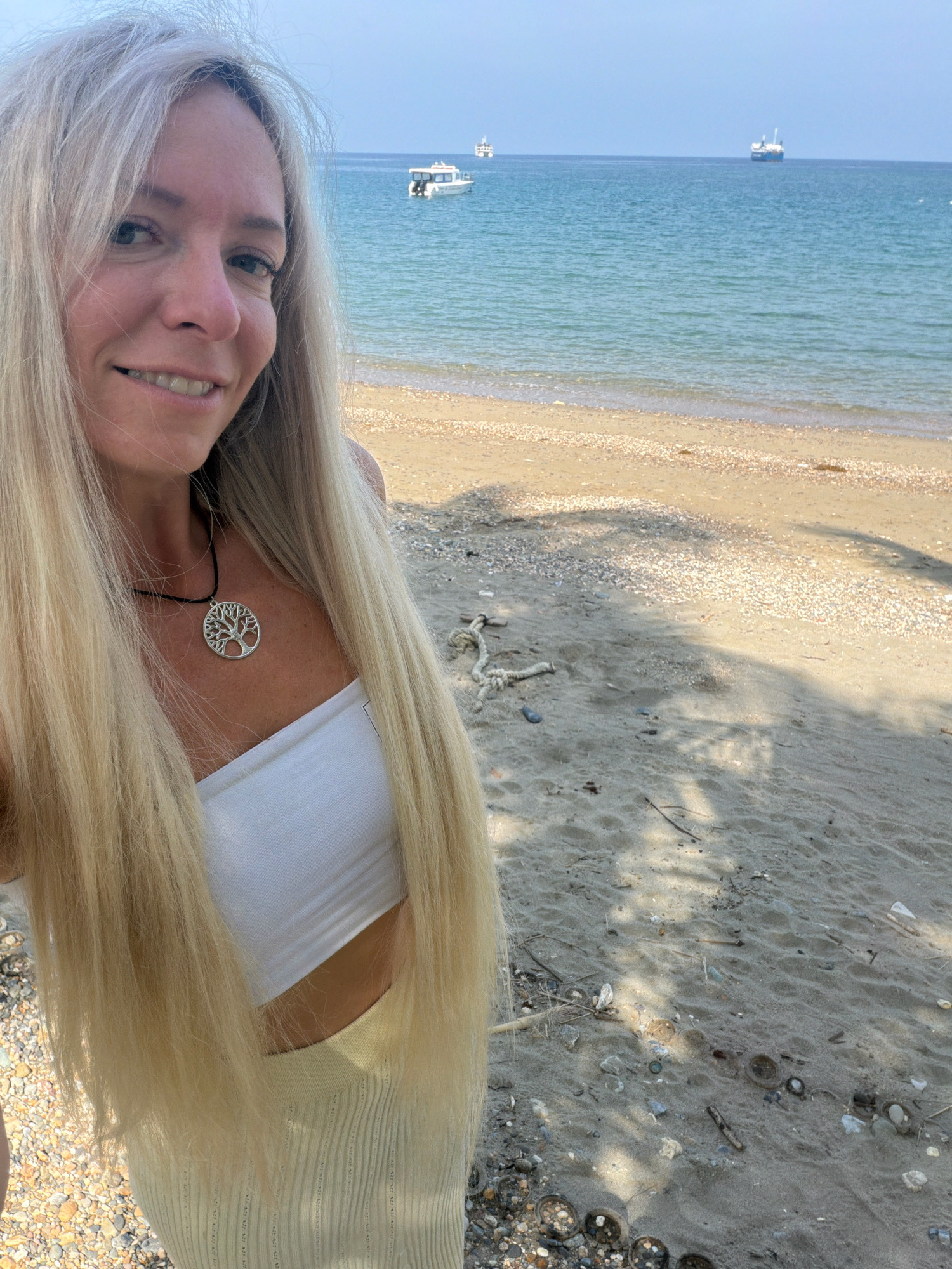 A blonde woman is standing on a sandy beach in Malay, Philippines. The woman is wearing a white tube top and a light-colored skirt. She has a necklace with a tree of life pendant. In the background, there is a body of water with a few boats visible. The sand is littered with shells and debris. The lighting suggests it is a sunny day.