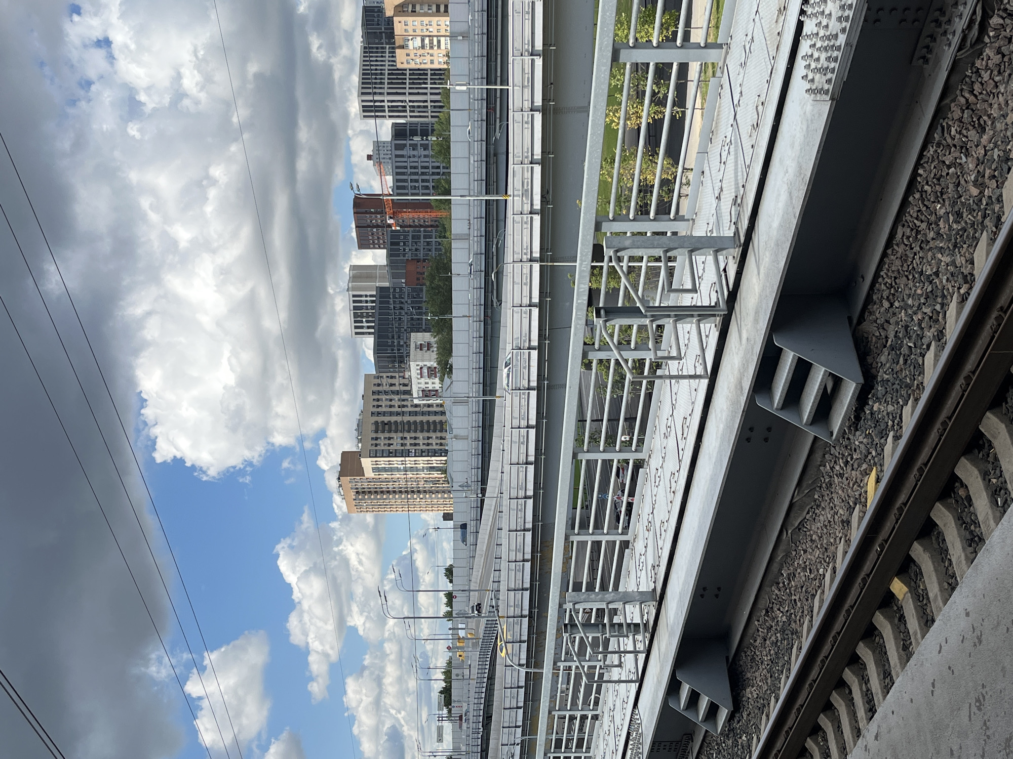 The image depicts a multi-level urban environment with an emphasis on infrastructure and city architecture under a partly cloudy sky. In the foreground, train tracks run horizontally, with ballast and sleepers clearly visible. Adjacent to the tracks is a raised metal platform with stairs, railings, and a metal grate surface.
Beyond the train platform, several elevated roadways and bridges are visible. Cars are seen driving on these roads, suggesting active traffic flow. The bridges have concrete barriers and railings, with streetlights lining some of the elevated sections.
In the background, a dense cluster of modern residential buildings rises against the sky. These buildings vary in height and architectural style, featuring a mix of light and dark facades, with some exhibiting distinctive geometric patterns. The architecture suggests a contemporary urban development. A single orange construction crane is visible amongst the buildings.
The sky is a mix of bright blue with scattered white cumulus clouds and darker, more ominous grey clouds, indicating dynamic weather conditions. The scene is characteristic of a modern, rapidly developing area within Moscow, Russia, possibly a transportation hub or a newly constructed residential district. There are no people visible in the image.