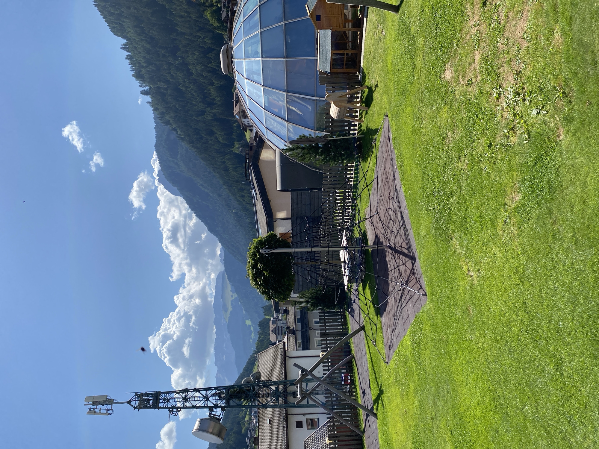 This image captures a sunny day in the town of Ortisei, Italy, showcasing a playground with a backdrop of lush green mountains. In the foreground, a vibrant green lawn surrounds a climbing net structure, its ropes forming a complex pattern. To the left, a wooden swing set with a red seat stands on a rubberized mat. Further to the left, a tall cell tower with multiple antennas dominates the sky. Beyond the playground, a series of houses with tiled roofs are visible, nestled against the slopes. A distinctive geodesic dome, with its blue, segmented glass panels, stands out amongst the buildings. The surrounding mountains are covered in dense green pine forests, reaching towards a bright blue sky dotted with fluffy white clouds. The overall scene conveys a peaceful, outdoor atmosphere in a mountainous setting.