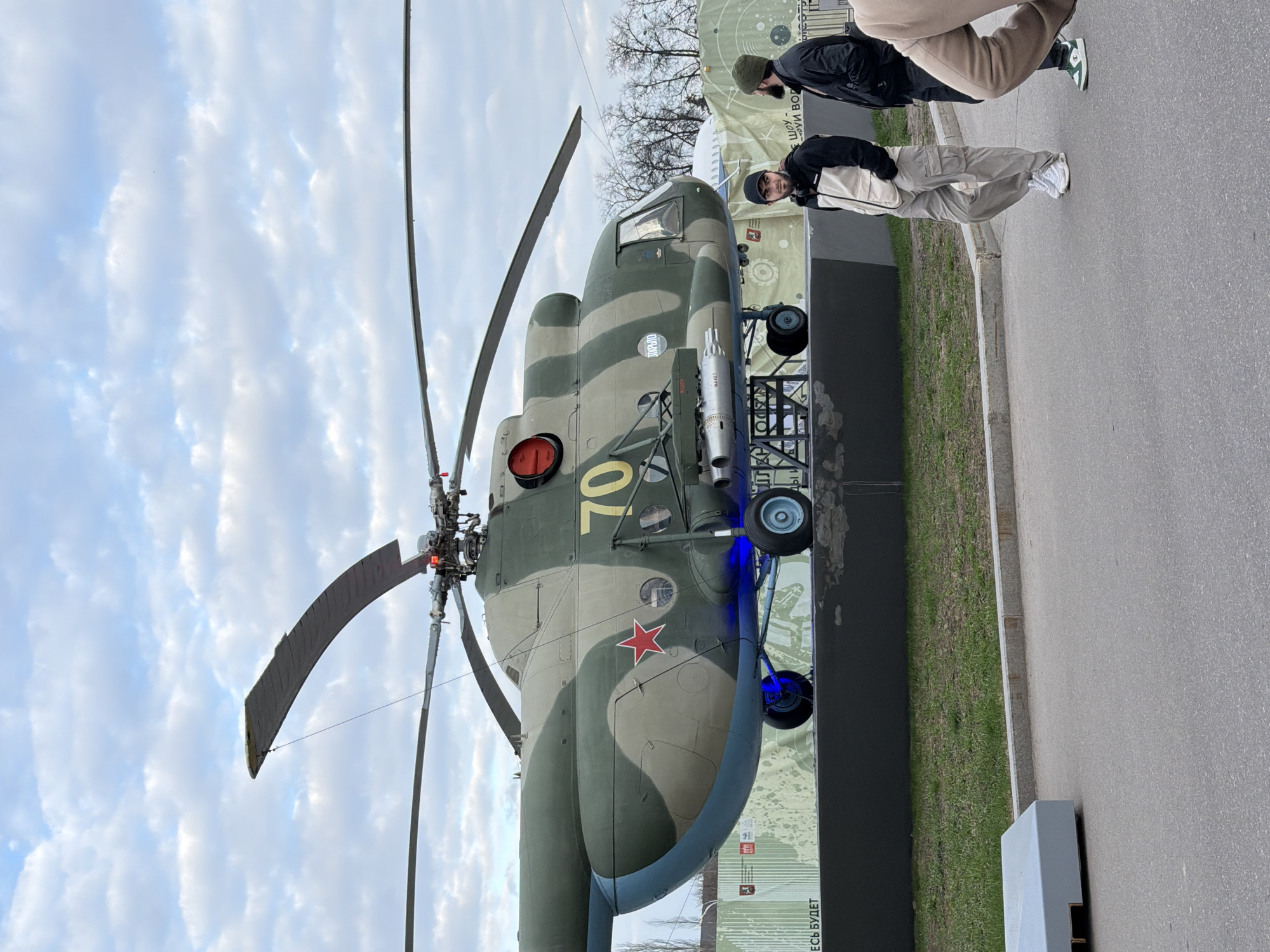 The image depicts a Mi-24 Hind attack helicopter on display in Moscow, Russia. The helicopter is painted in a camouflage pattern of green and brown with the number "70" painted in yellow. It has a red star emblem on the fuselage and is equipped with missiles. The background includes a cloudy sky, trees, a display stand with text in Russian, and people walking by. The ground is paved.