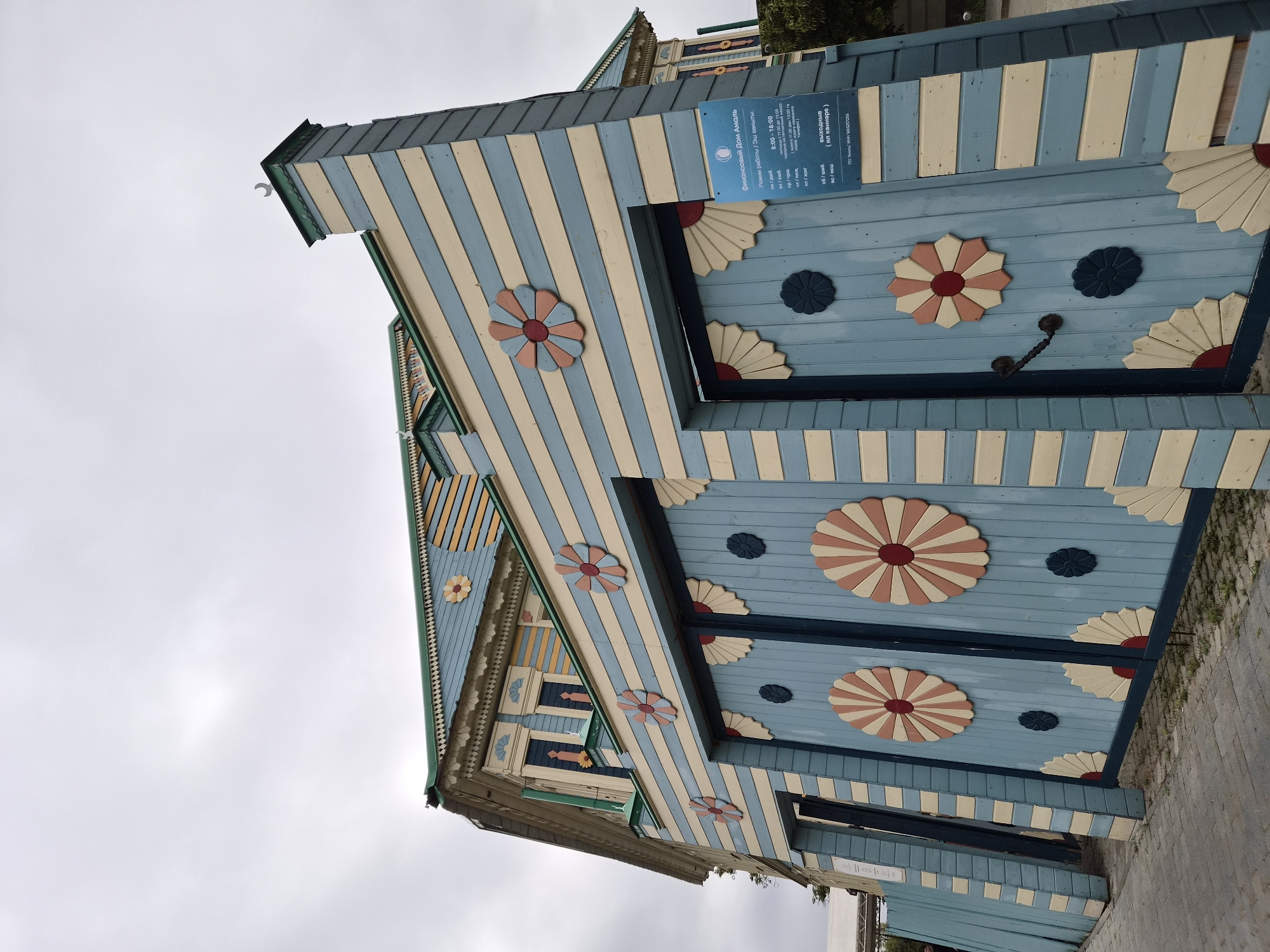 The image shows a vibrantly painted wooden gate and the side of a building in Kazan, Russia. The gate is painted in alternating horizontal stripes of light blue and cream, with decorative floral motifs in red, pink, and cream. The building also features similar striped painting and ornamentation. A sign with text in Russian is affixed to the right side of the gate. The sky is overcast, and the ground in front of the gate is paved with cobblestones. The overall style of the architecture and decoration suggests a traditional or folk art influence.